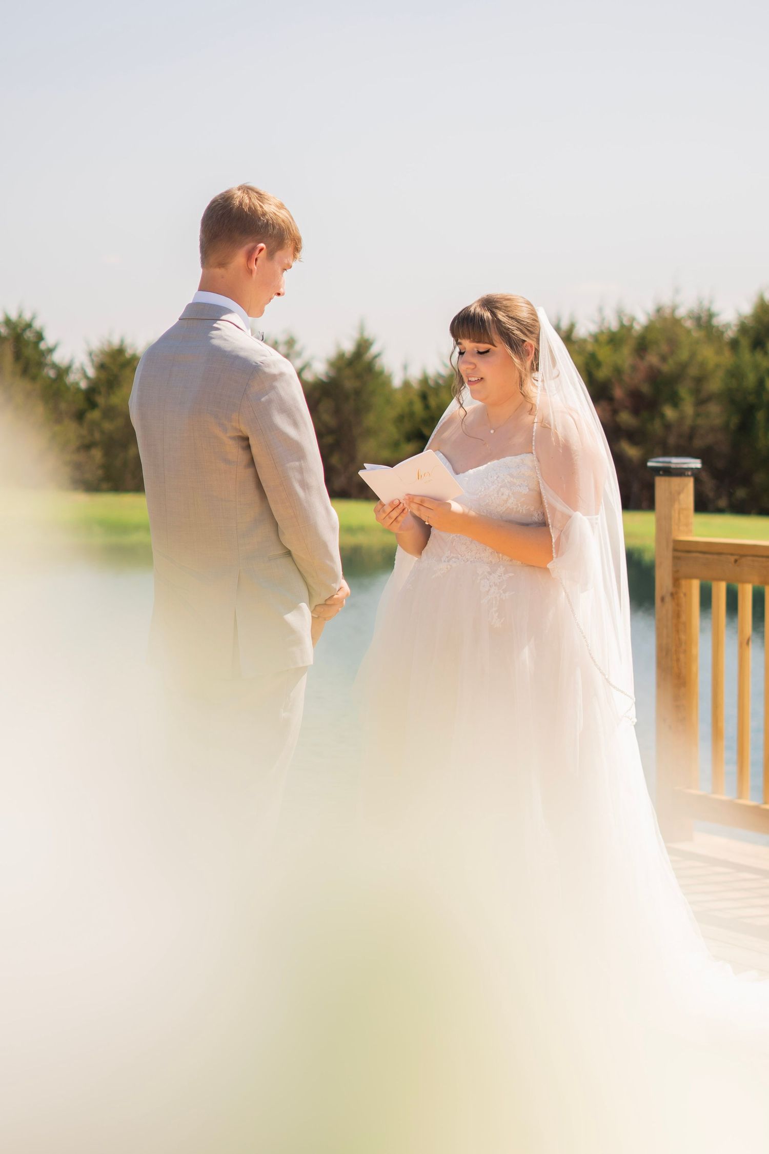 Bride reads vows to groom on outdoor deck in bright sunlight during intimate wedding ceremony.