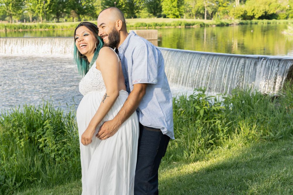 Couple embracing by waterfall in sunlit garden setting during maternity photoshoot.