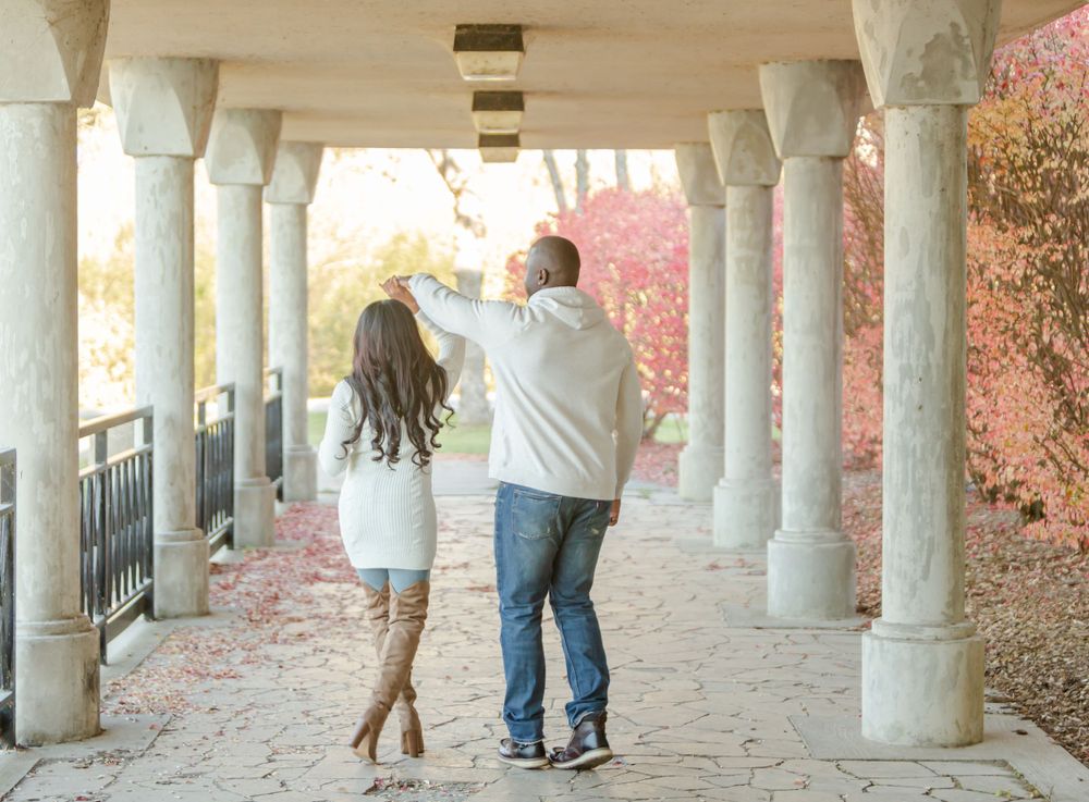 Couple walking together under colonnade with autumn foliage in background.