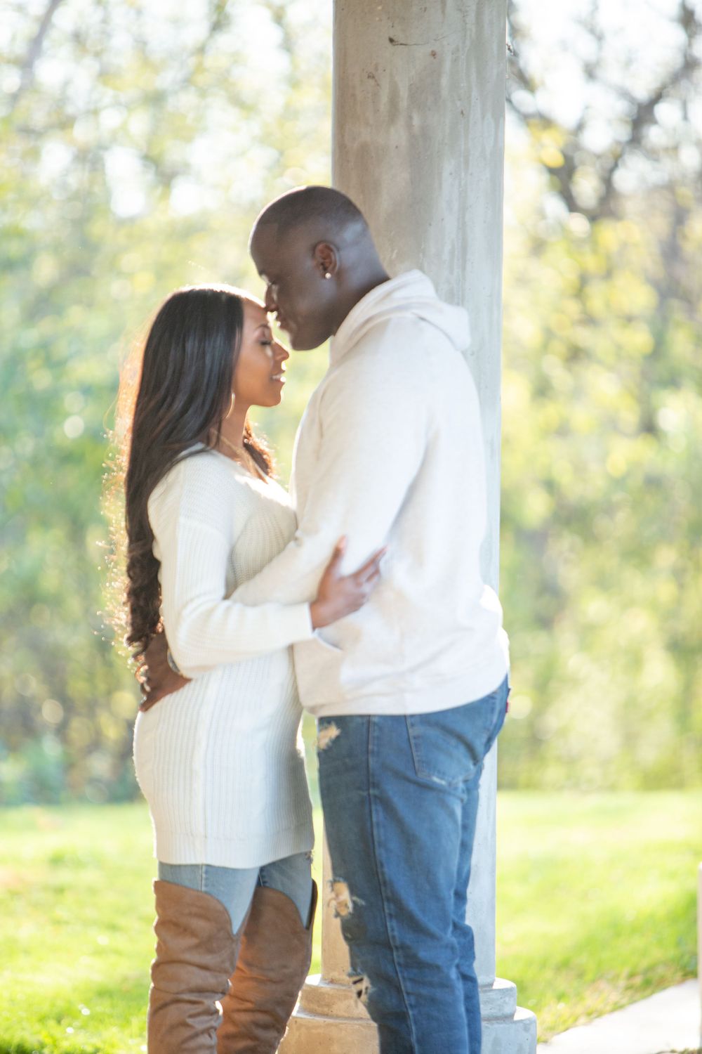 Couple in white sweaters sharing tender moment by white columns with greenery in background.