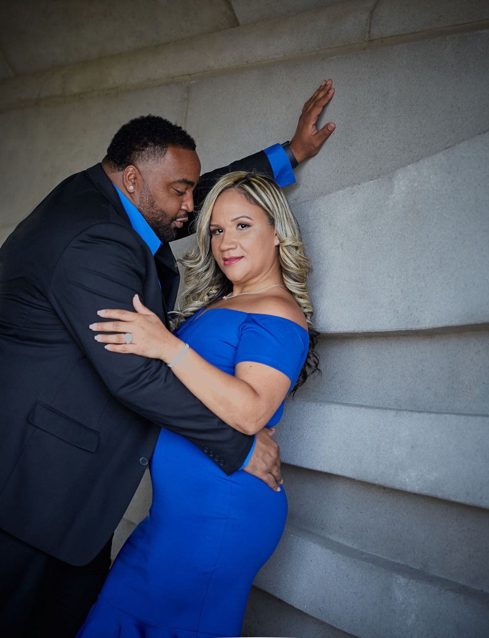 Romantic couple in formal wear posing against textured wall with dramatic lighting.