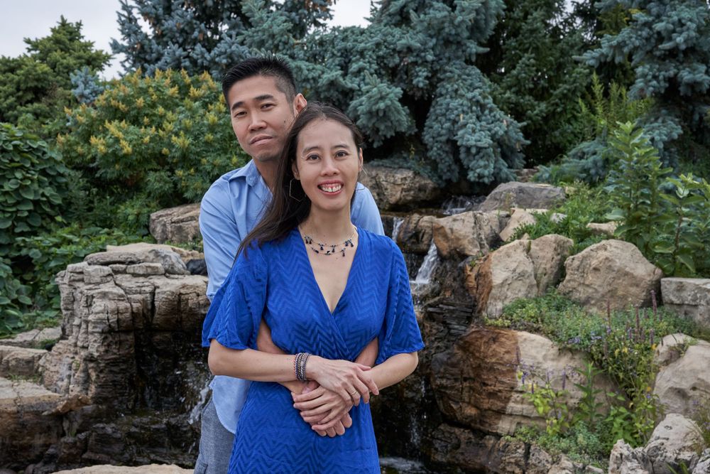 Couple in coordinated blue outfits posing by decorative garden pond with evergreens.