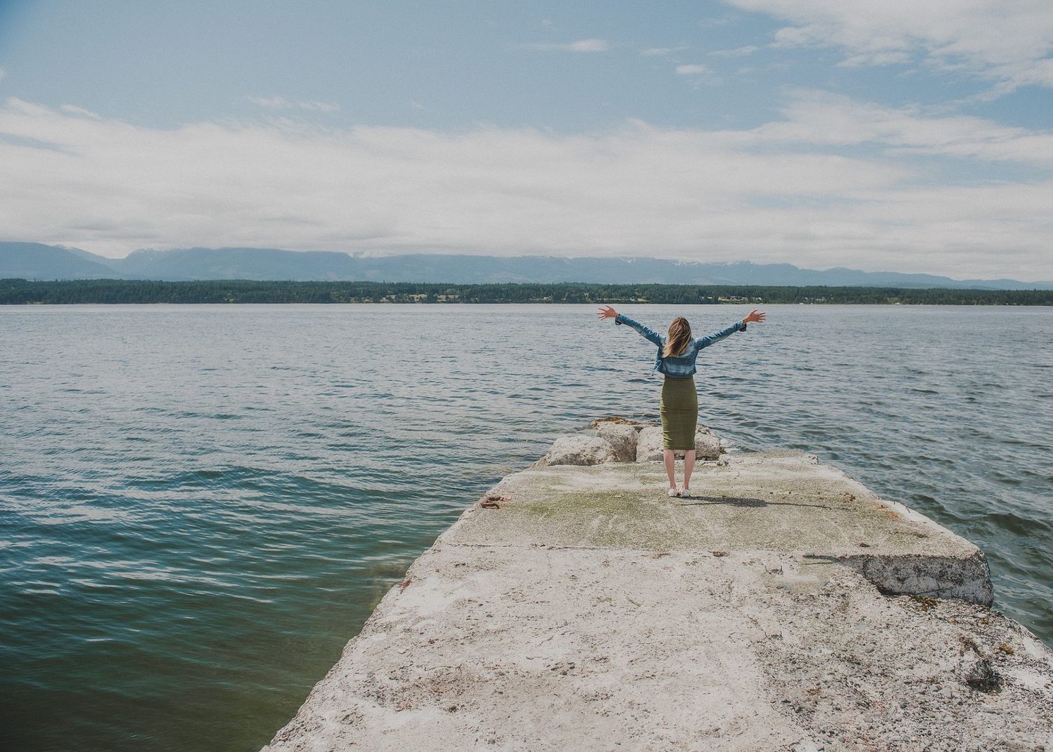 Silhouette of person standing with arms raised at the end of a concrete pier on a lake.