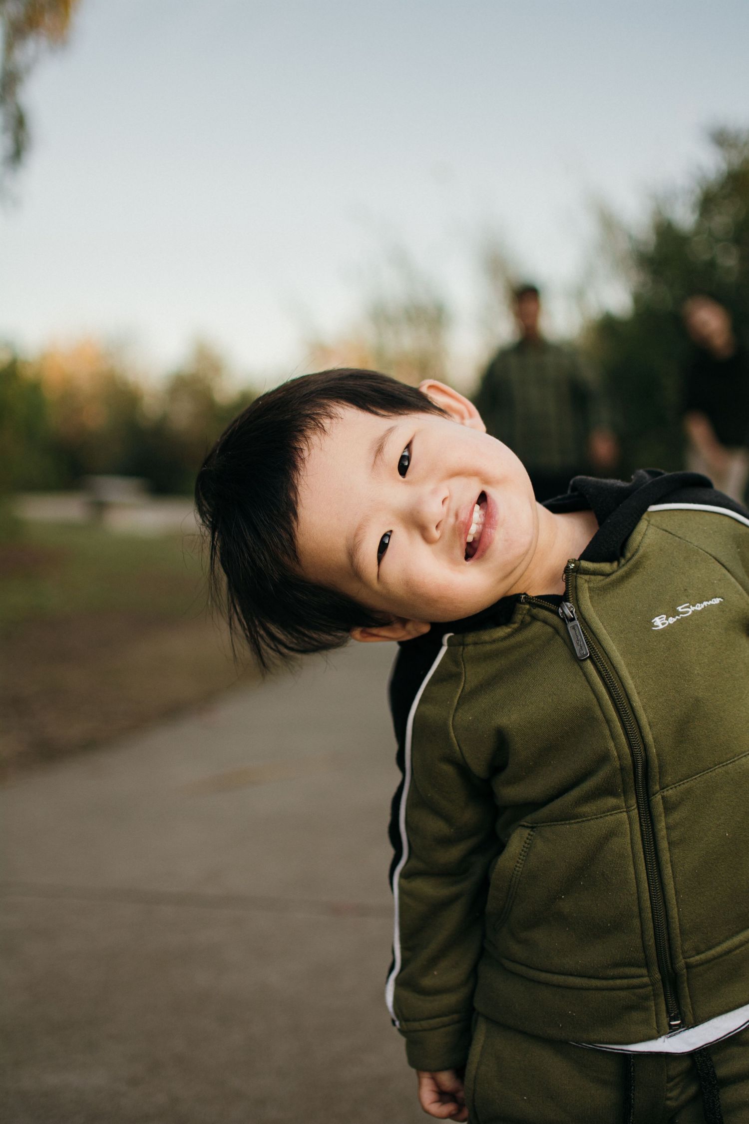 A candid outdoor portrait of a joyful young person wearing an olive green zip-up jacket.
