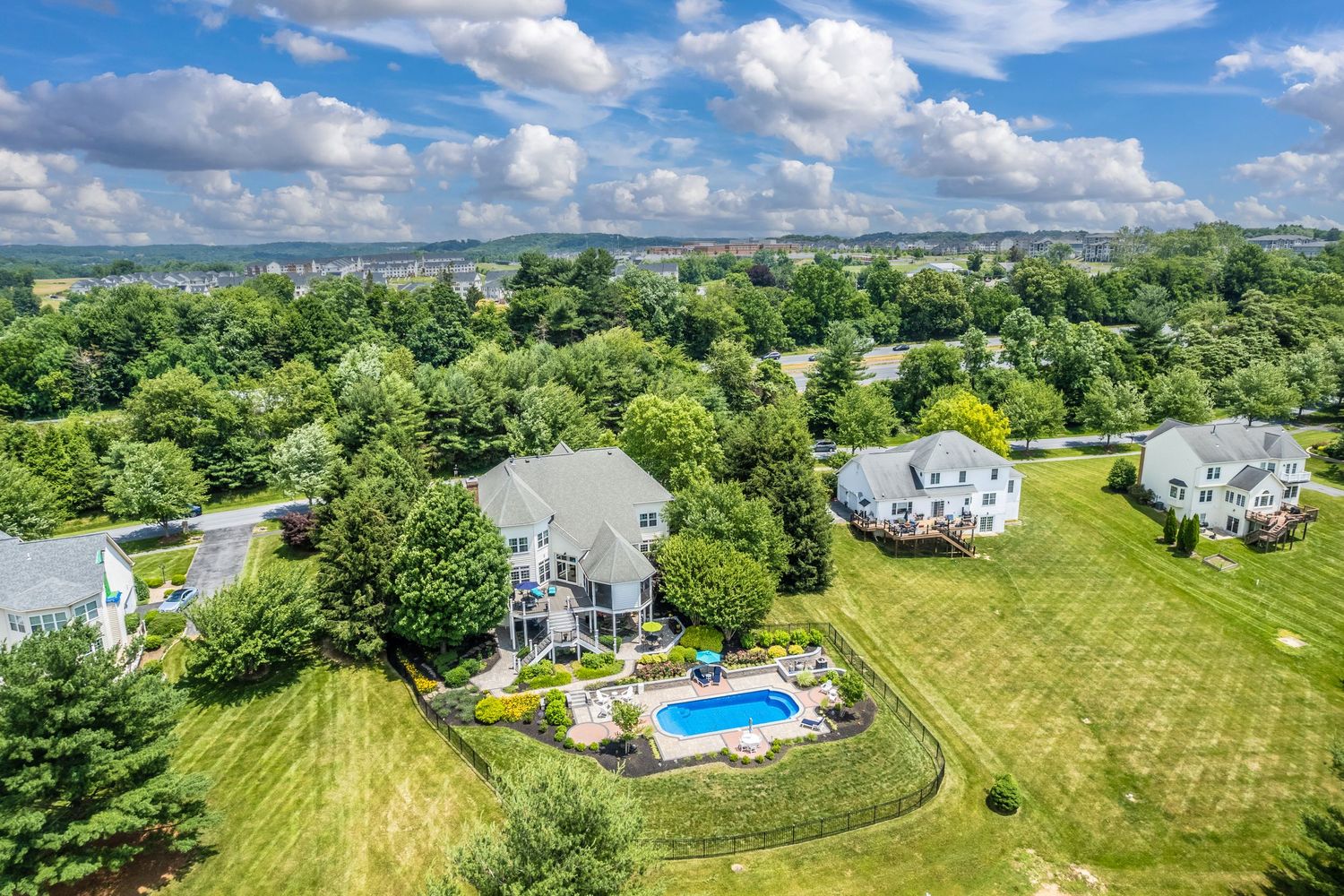 Aerial view of suburban homes with a backyard swimming pool surrounded by lush green trees under puffy white clouds.
