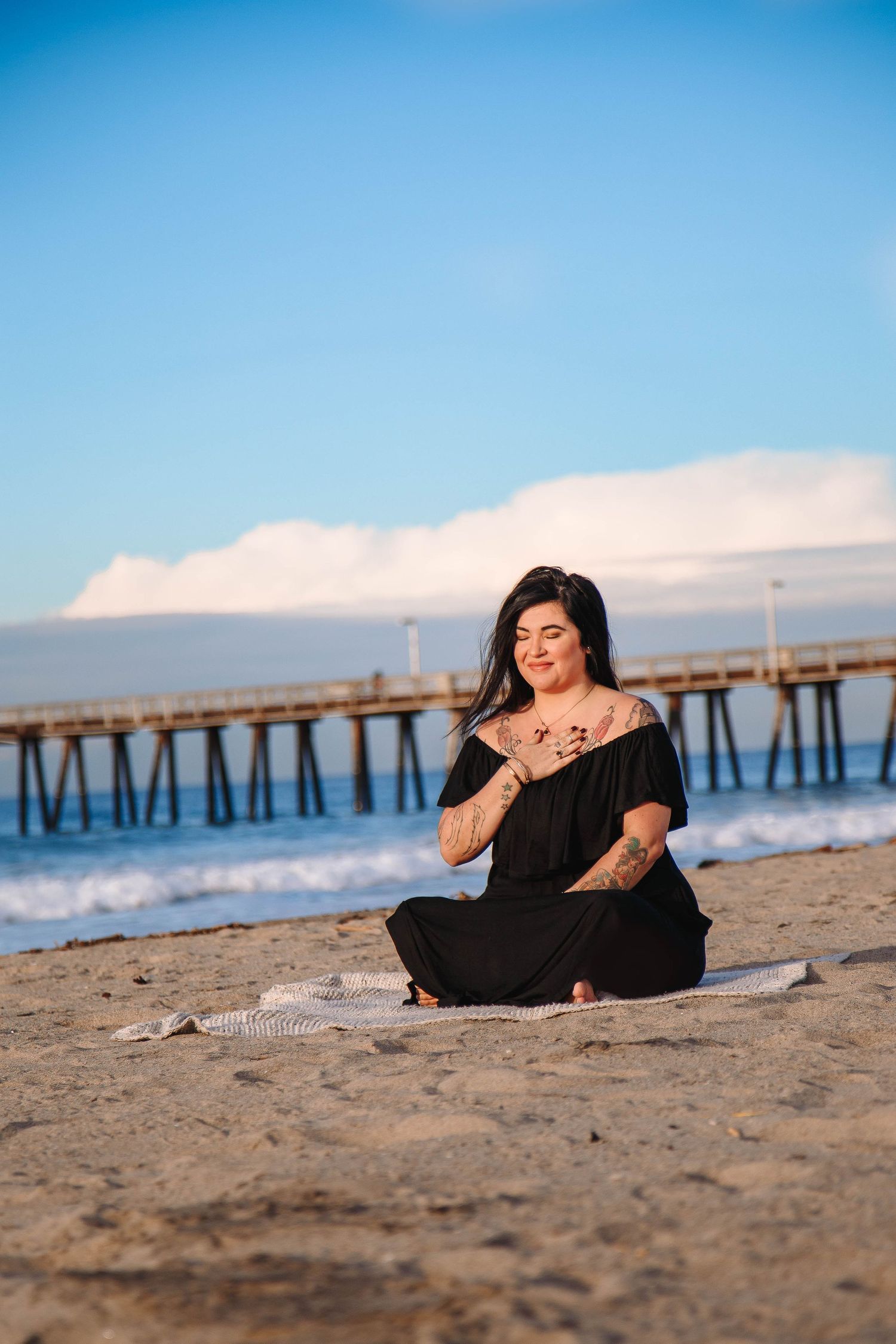 women in a black dress siting and meditating on the beach with the pier behind her
