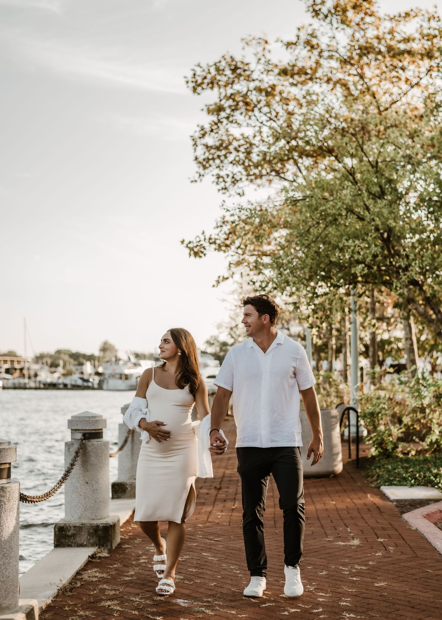 Couple strolling along a waterfront boardwalk at sunset with boats in the background.