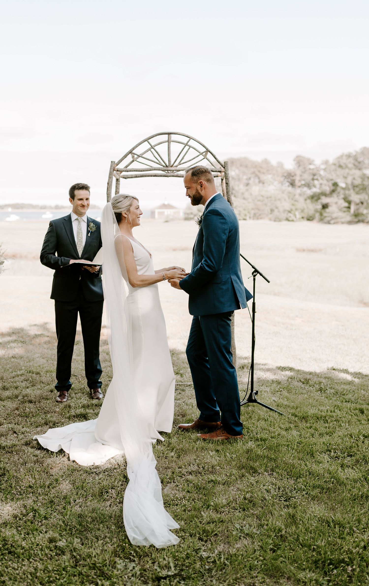 A romantic wedding ceremony takes place under a rustic arch on a grassy hillside.