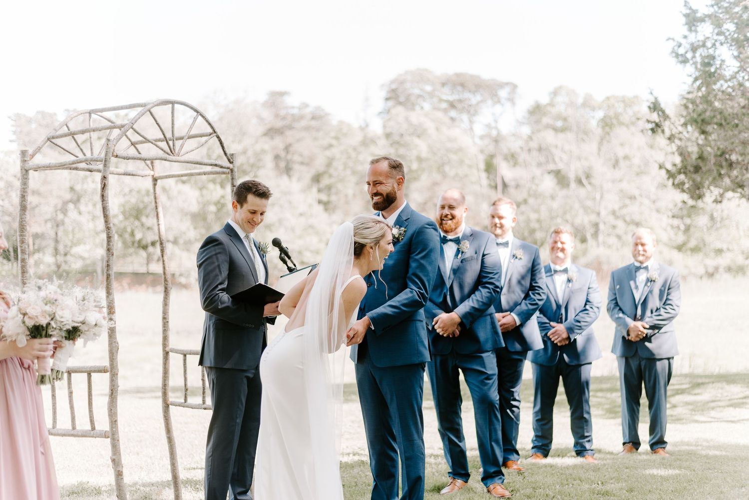 Wedding ceremony setup with rustic arch and groomsmen in navy suits.