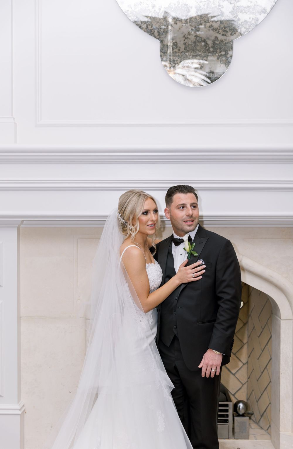 Couple posing by white fireplace in elegant wedding attire.