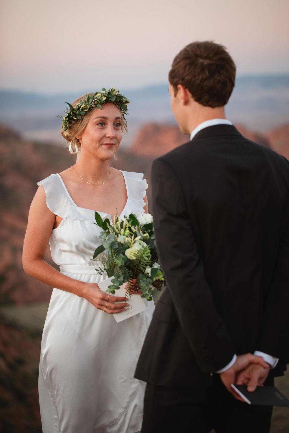 Bride in white dress and flower crown smiles at groom during red rock desert wedding ceremony.