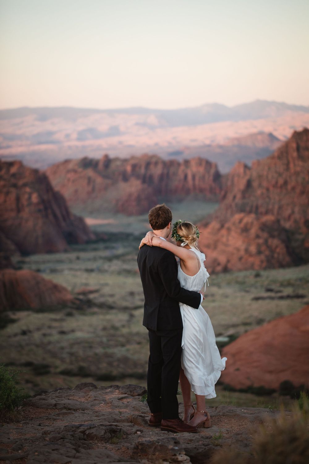 Bride and groom stand embracing overlooking breathtaking red rock canyon valley at sunset.
