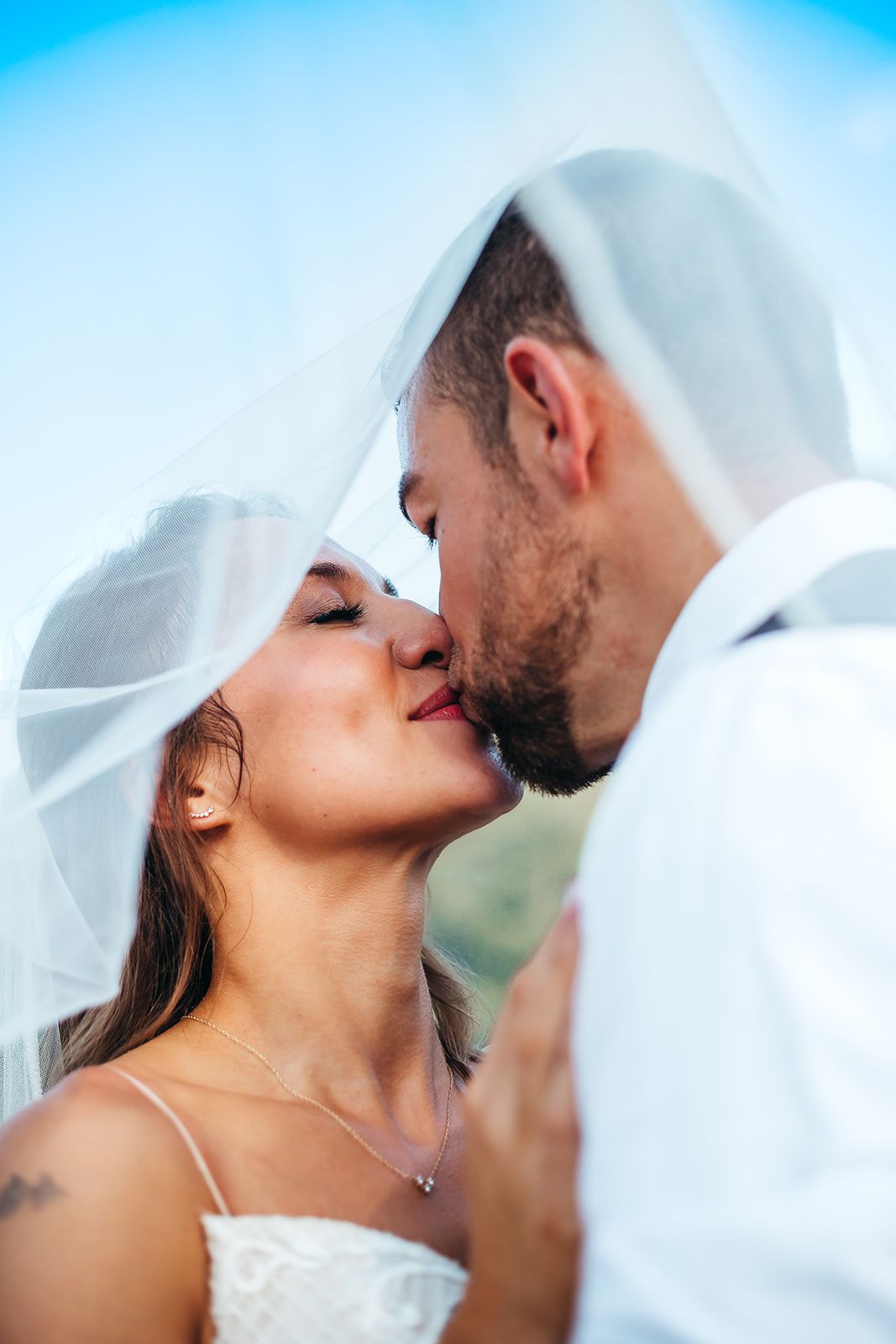 Romantic wedding couple sharing a tender moment under flowing white veil against blue sky.