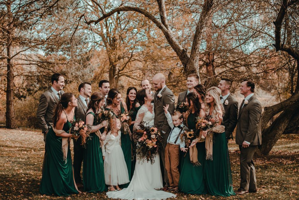 Wedding party in emerald green and brown attire celebrating among fall foliage outdoors.