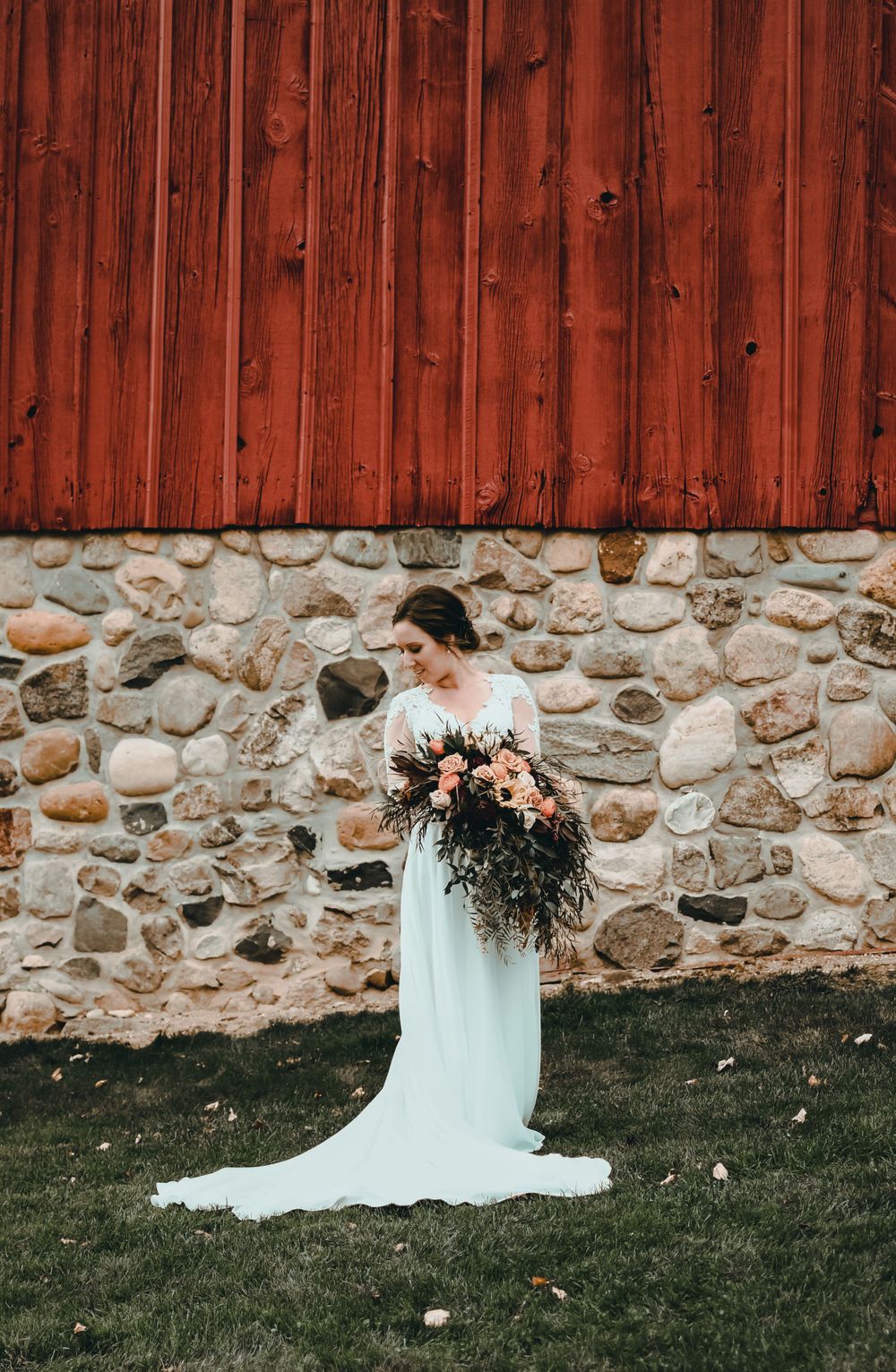 Bride in white gown holding lush bouquet standing before a rustic stone and red barn wall.
