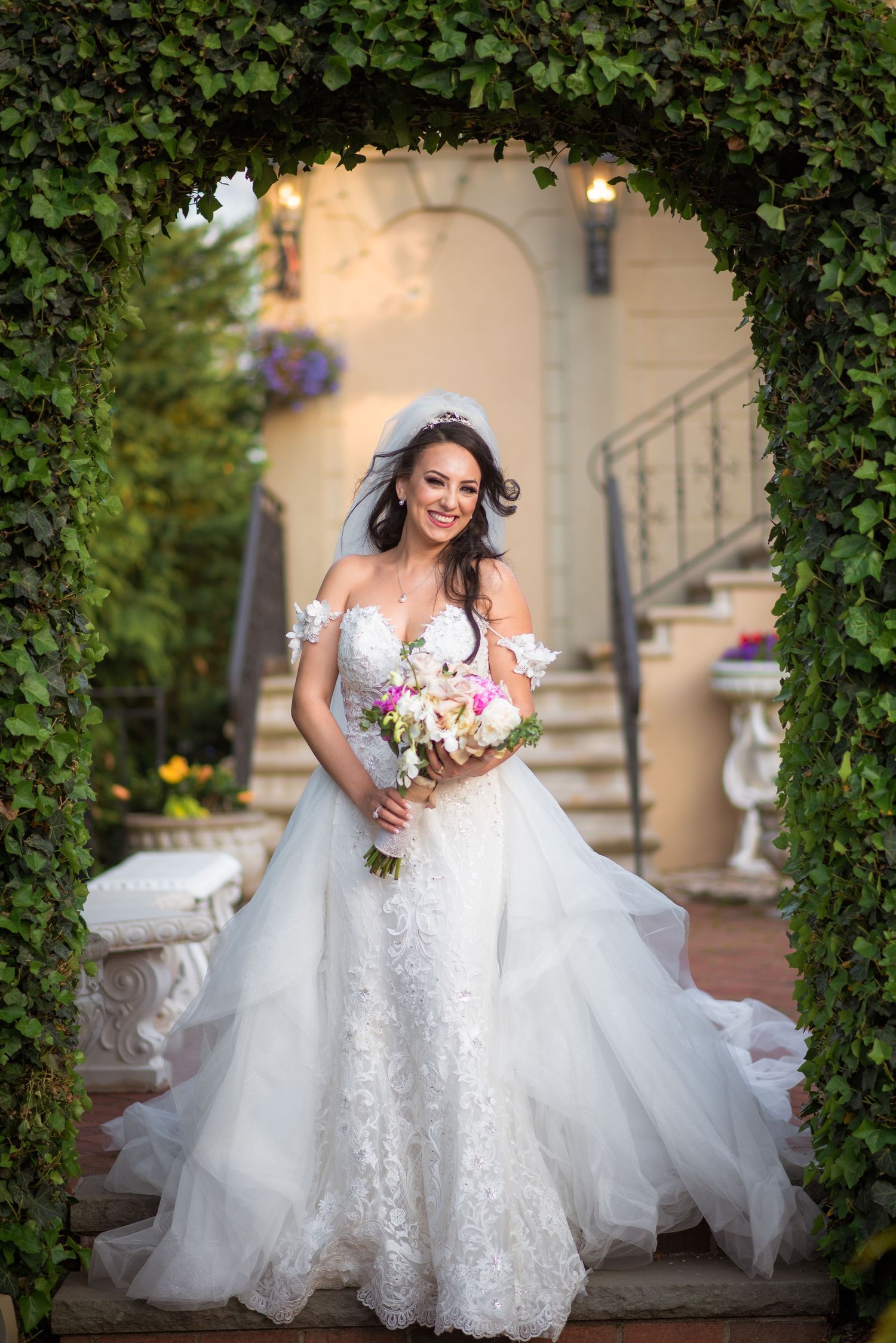 Elegant bride in off-shoulder lace gown holds bouquet under a lush green garden archway.