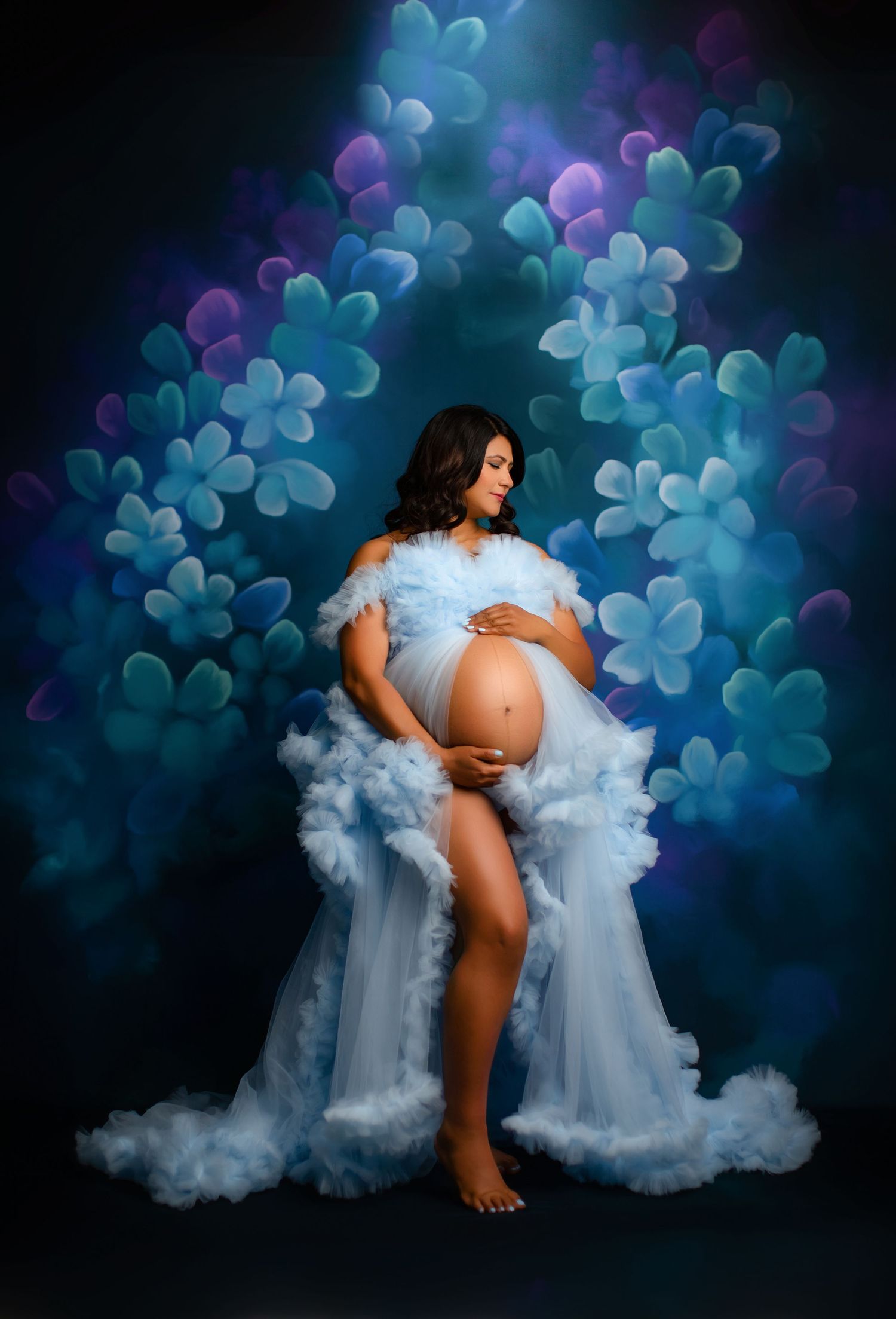 Dreamy maternity photo with flowing blue dress against dark floral backdrop.