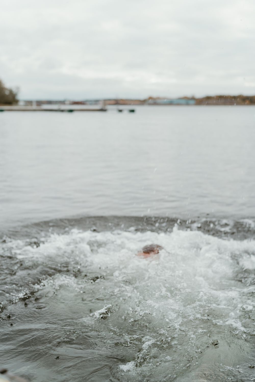Splashing water creates turbulent waves on a calm lake with buildings visible in the distant shoreline.