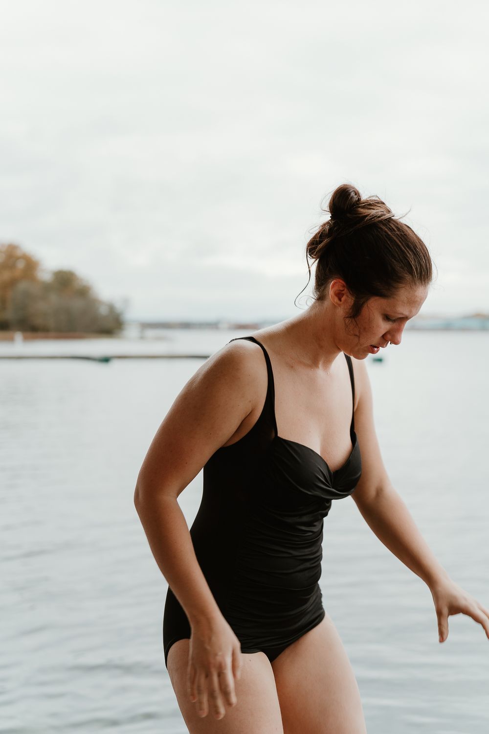 A swimmer in a black bathing suit stands at the edge of a lake on an overcast day.