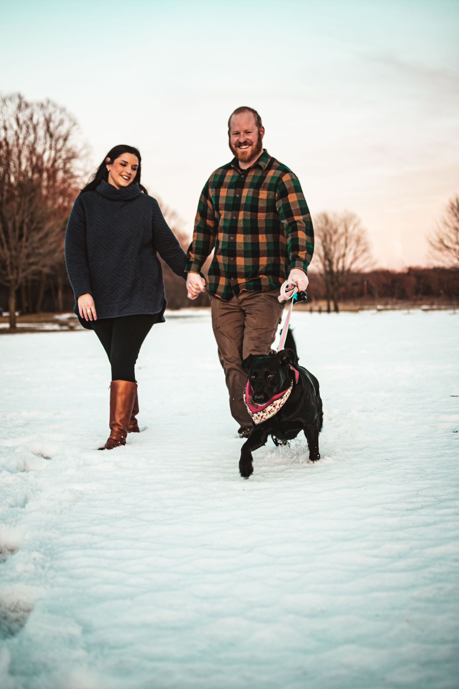 Couple walking their black dog through a snowy landscape at dusk.