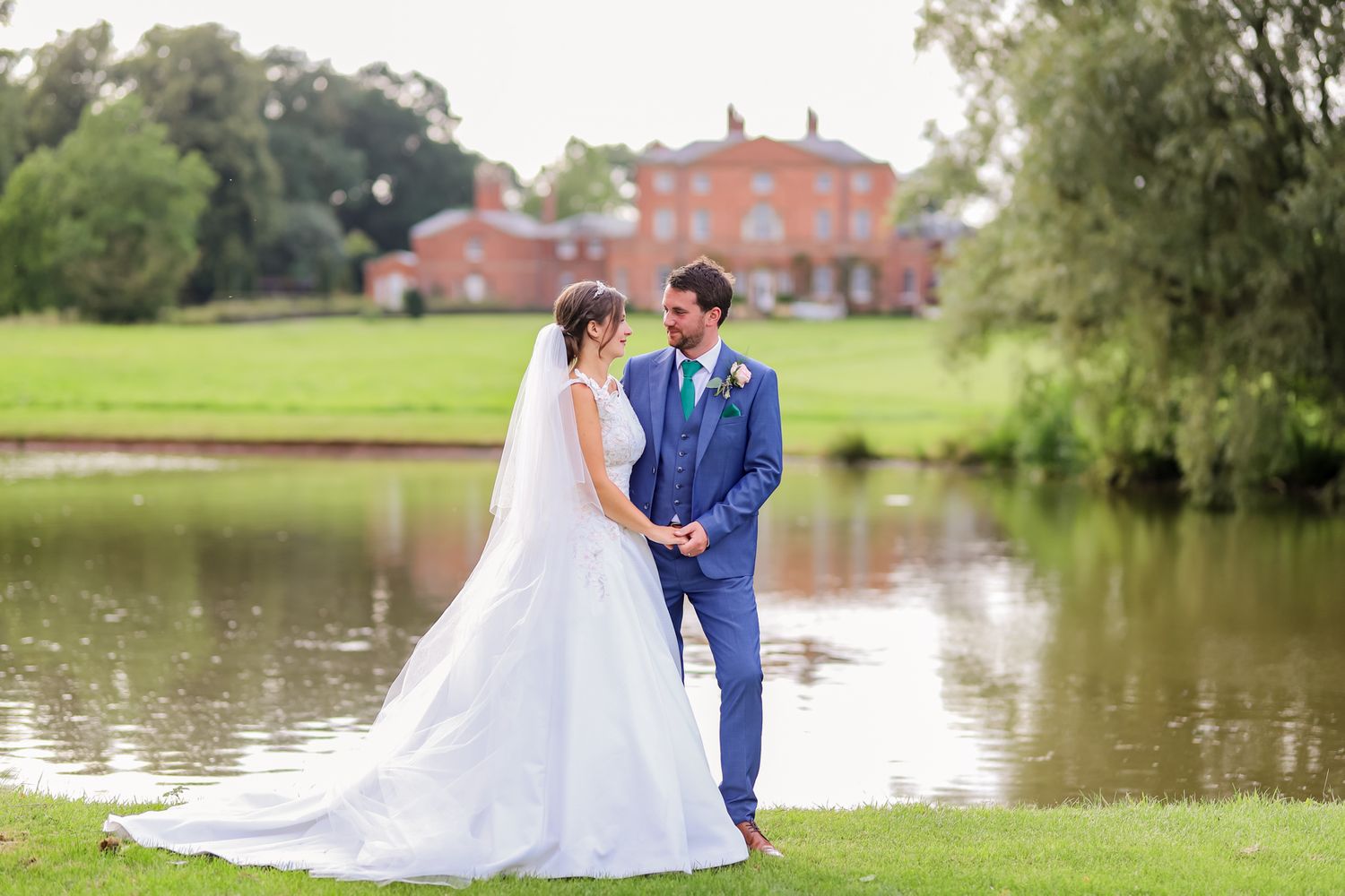 A couple poses for romantic photos by a lake with historic buildings in the background.
