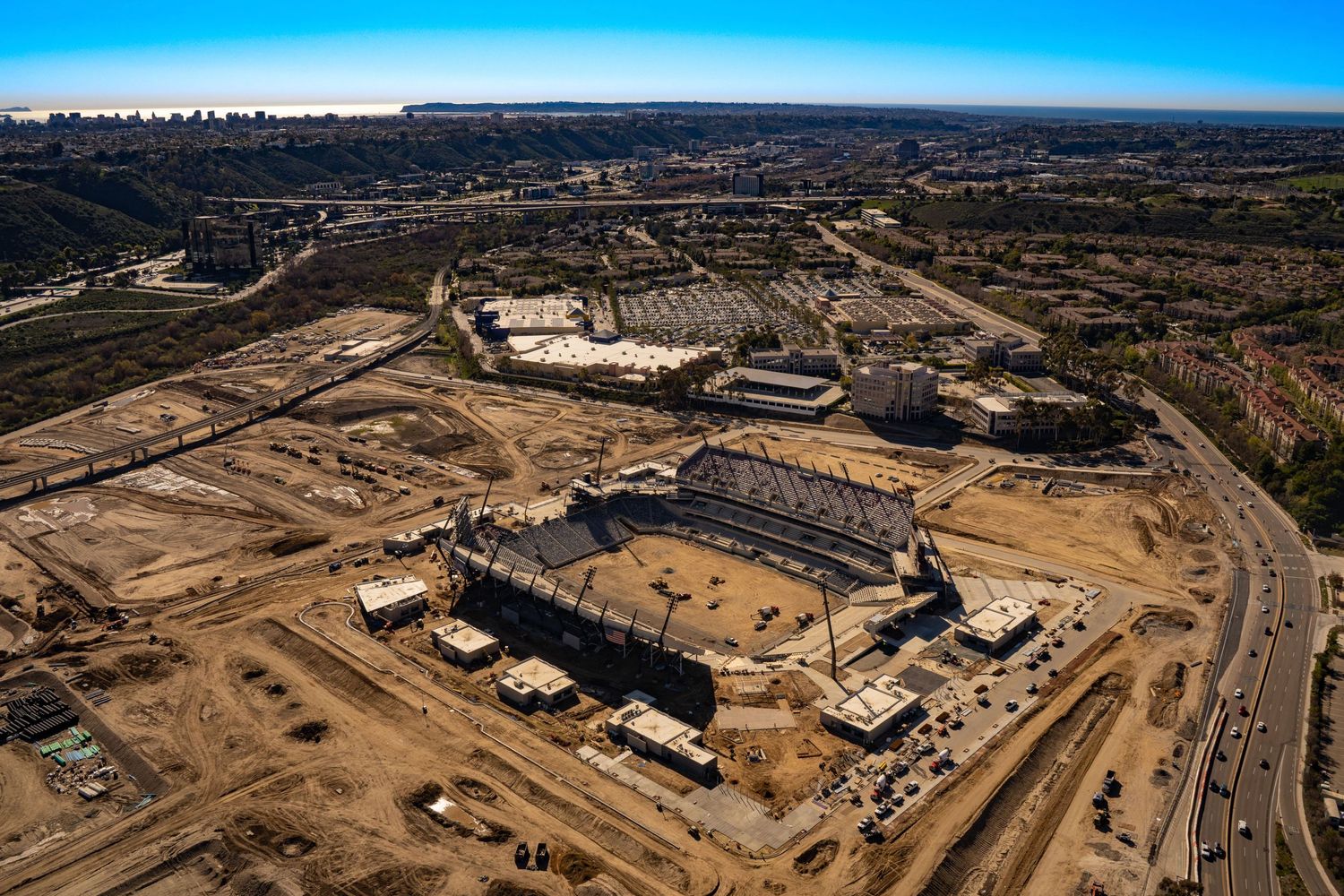 Aerial view of a large sports stadium under construction, showing grandstand structures surrounded by graded dirt lots, construction trailer