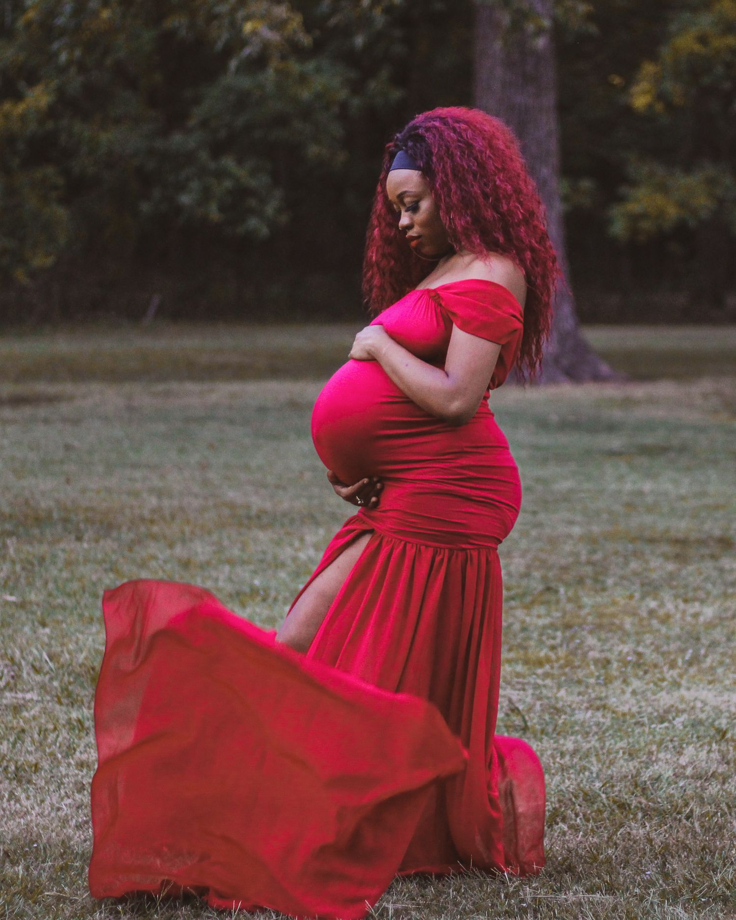 Elegant maternity photo of a figure in a flowing red dress standing in a grassy outdoor setting at dusk.