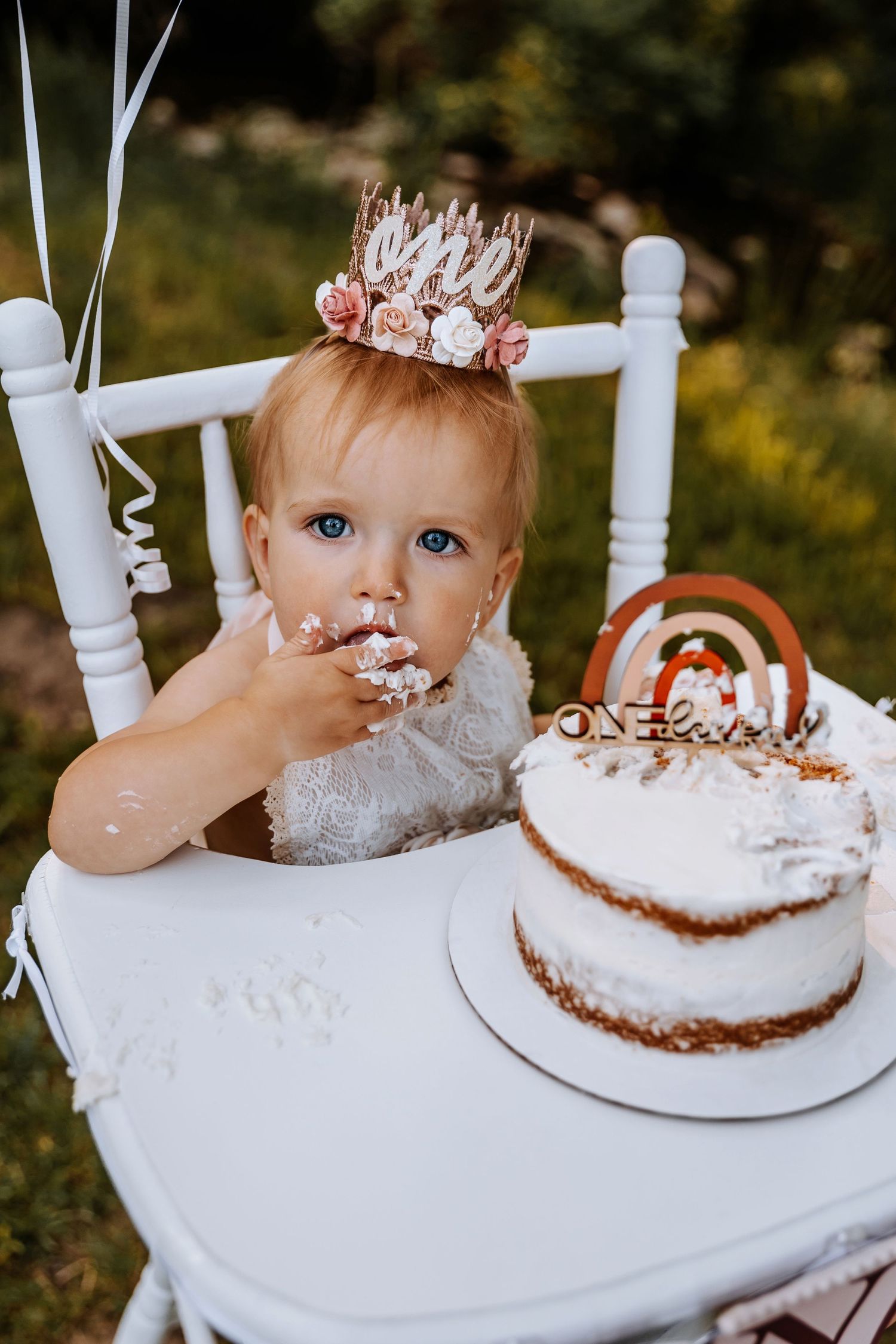 Baby in crown enjoying first birthday cake at white table in outdoor setting.