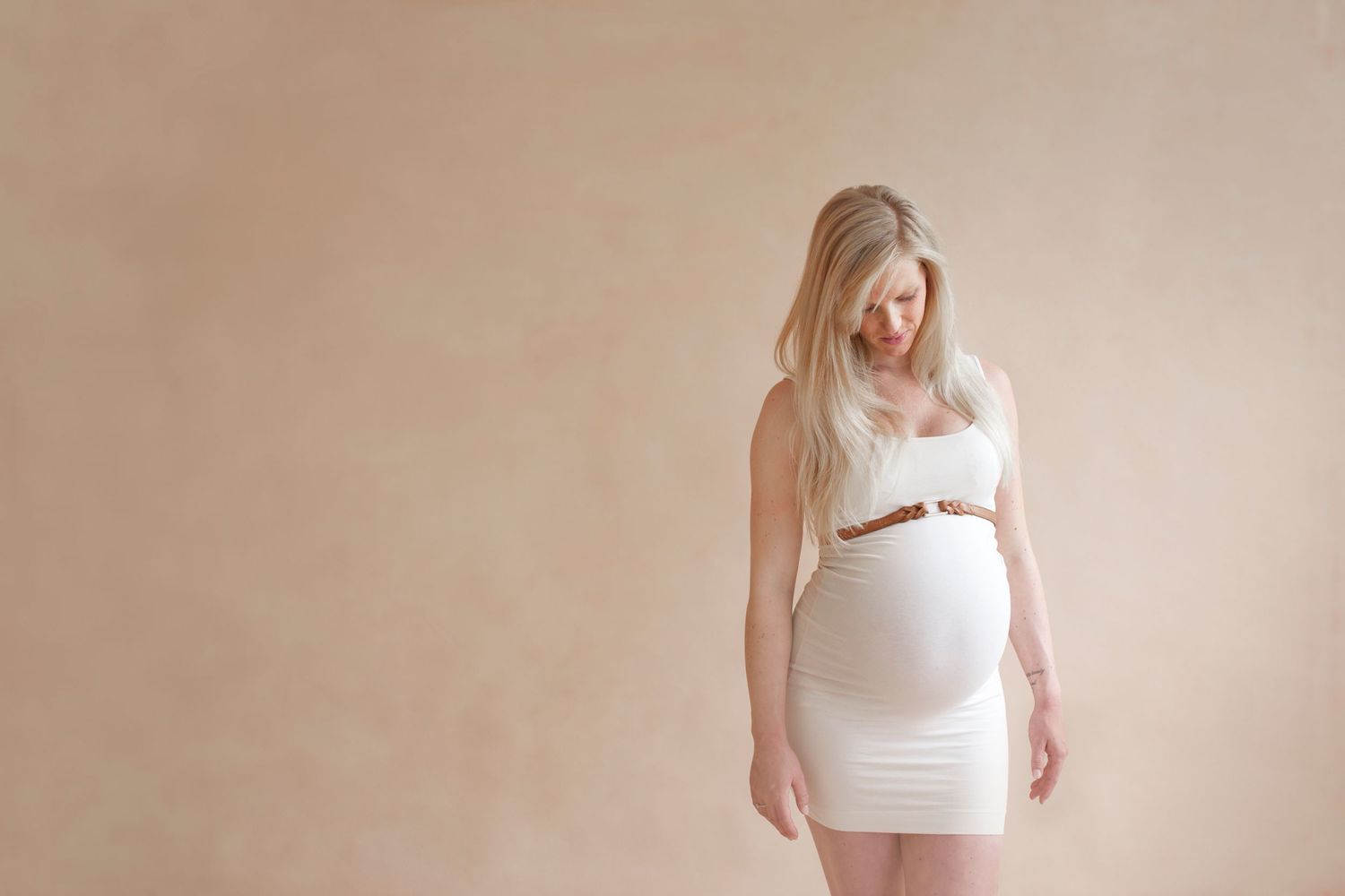 Expectant mother in white dress with brown belt poses against beige backdrop.