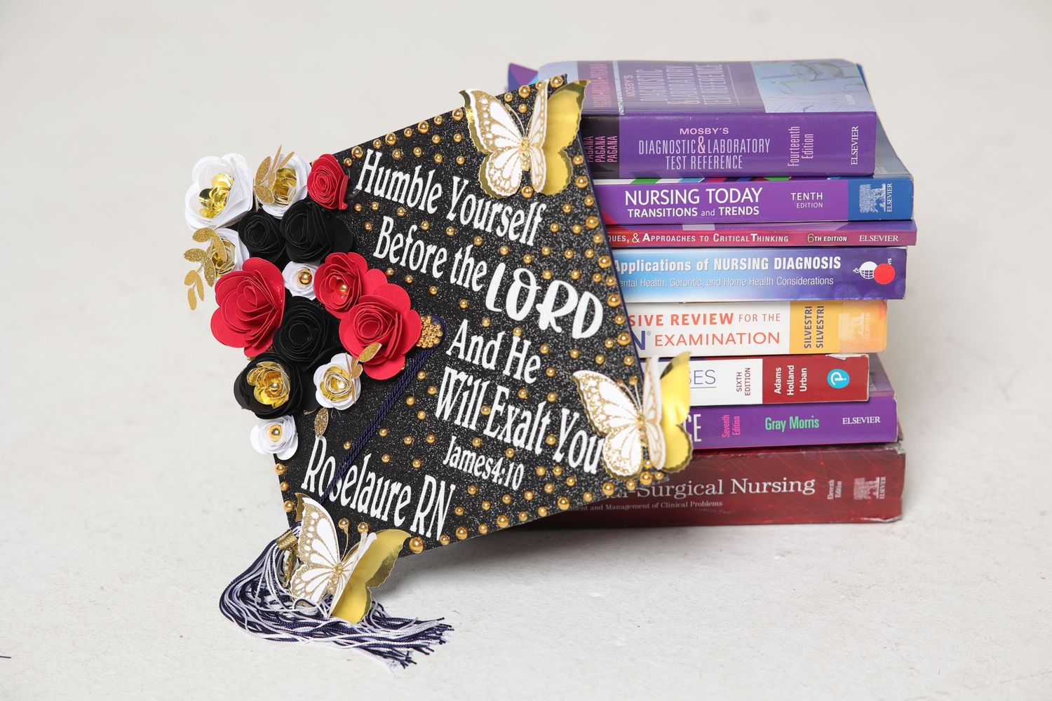 Decorated graduation cap reads Humble Yourself Before the Lord beside nursing textbooks stacked together.