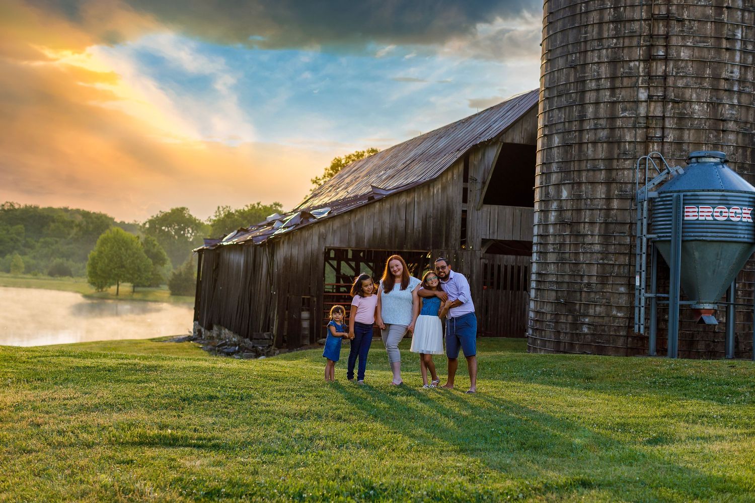 People gather outside a rustic barn at sunset with a grain silo and golden sky in the rural countryside.