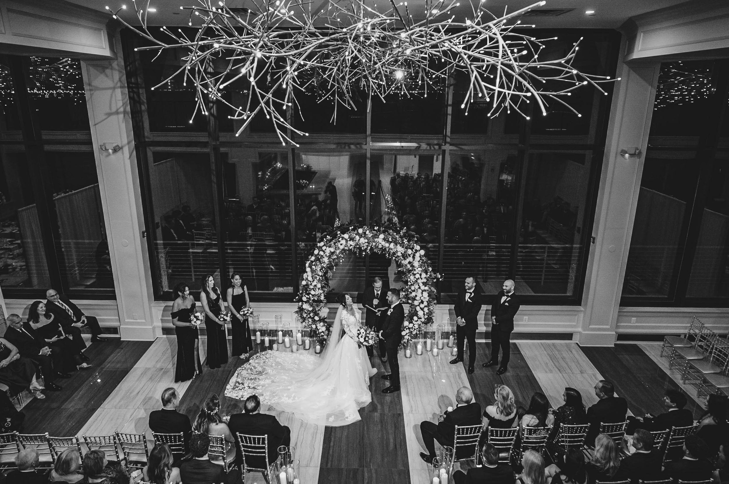 Wedding ceremony setup with floral arch and chandelier in modern venue with floor-to-ceiling windows.