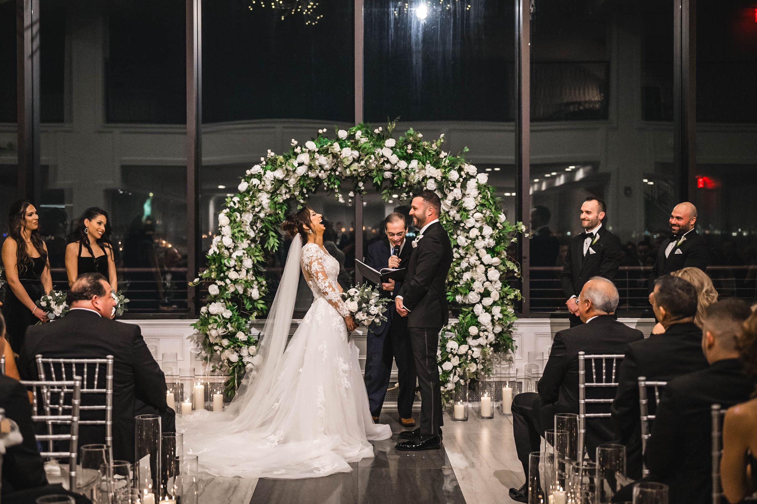 Wedding ceremony under floral arch with guests seated in elegant white chairs.