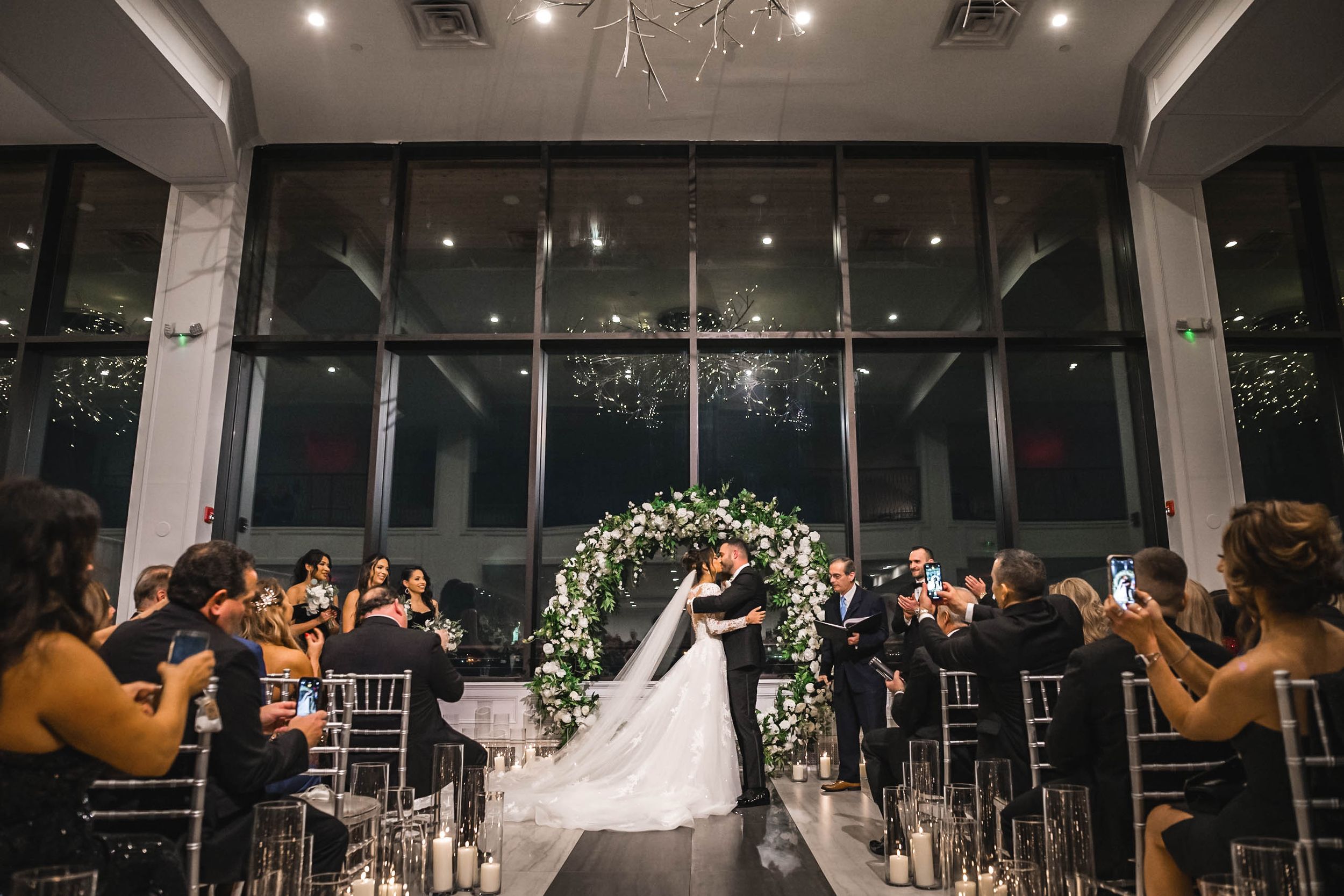 Wide angle view of wedding ceremony in modern venue with large windows and floral arch.