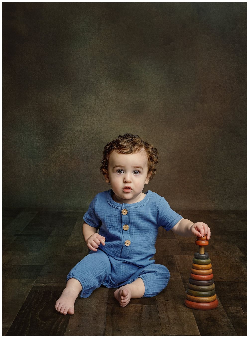 curly haired little boy in blue linen outfit on vintage wood floor and dark background posed with antique stacking toy.
