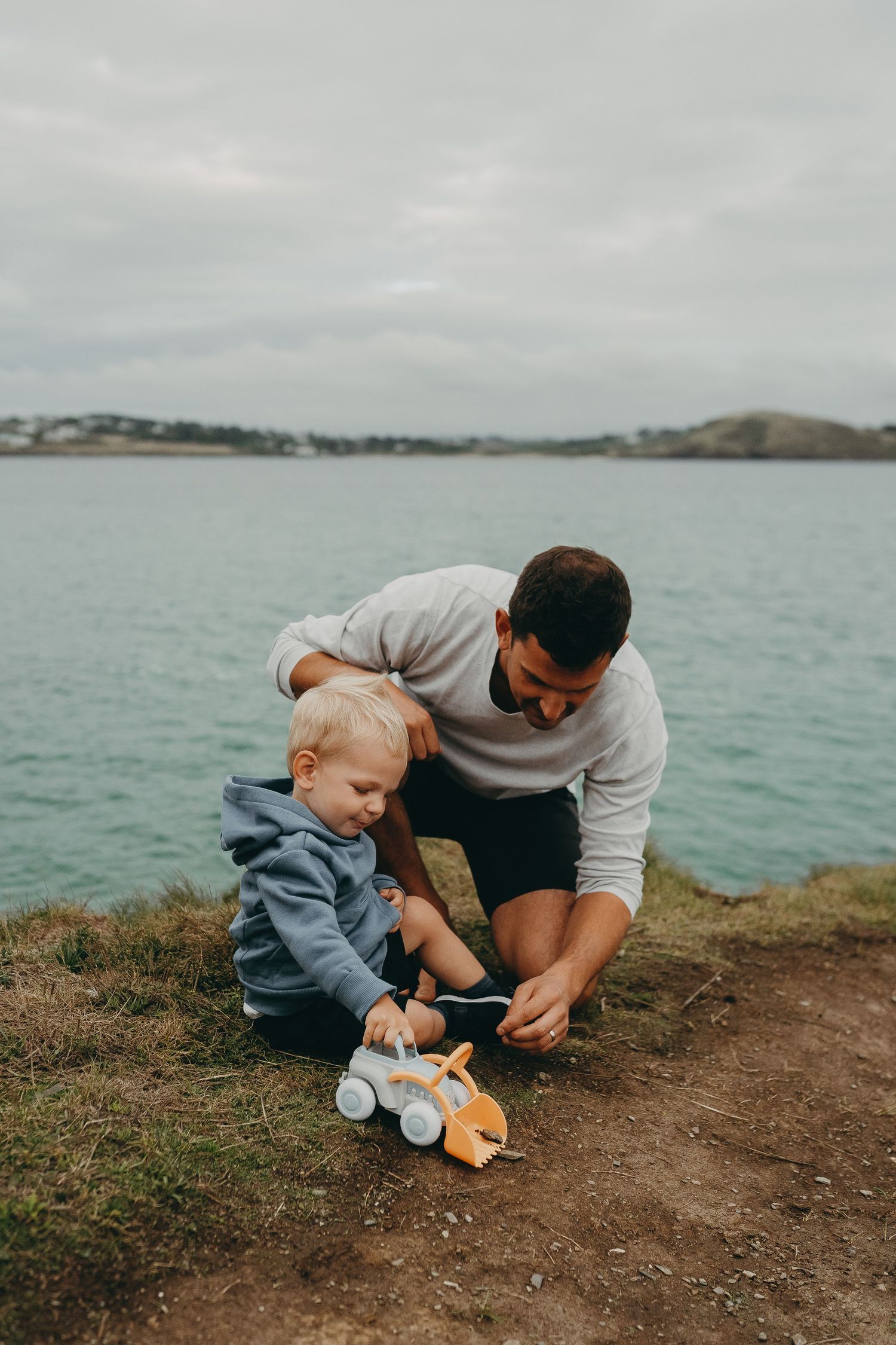 Parent and toddler playing with toy on coastal cliff overlooking blue waters.