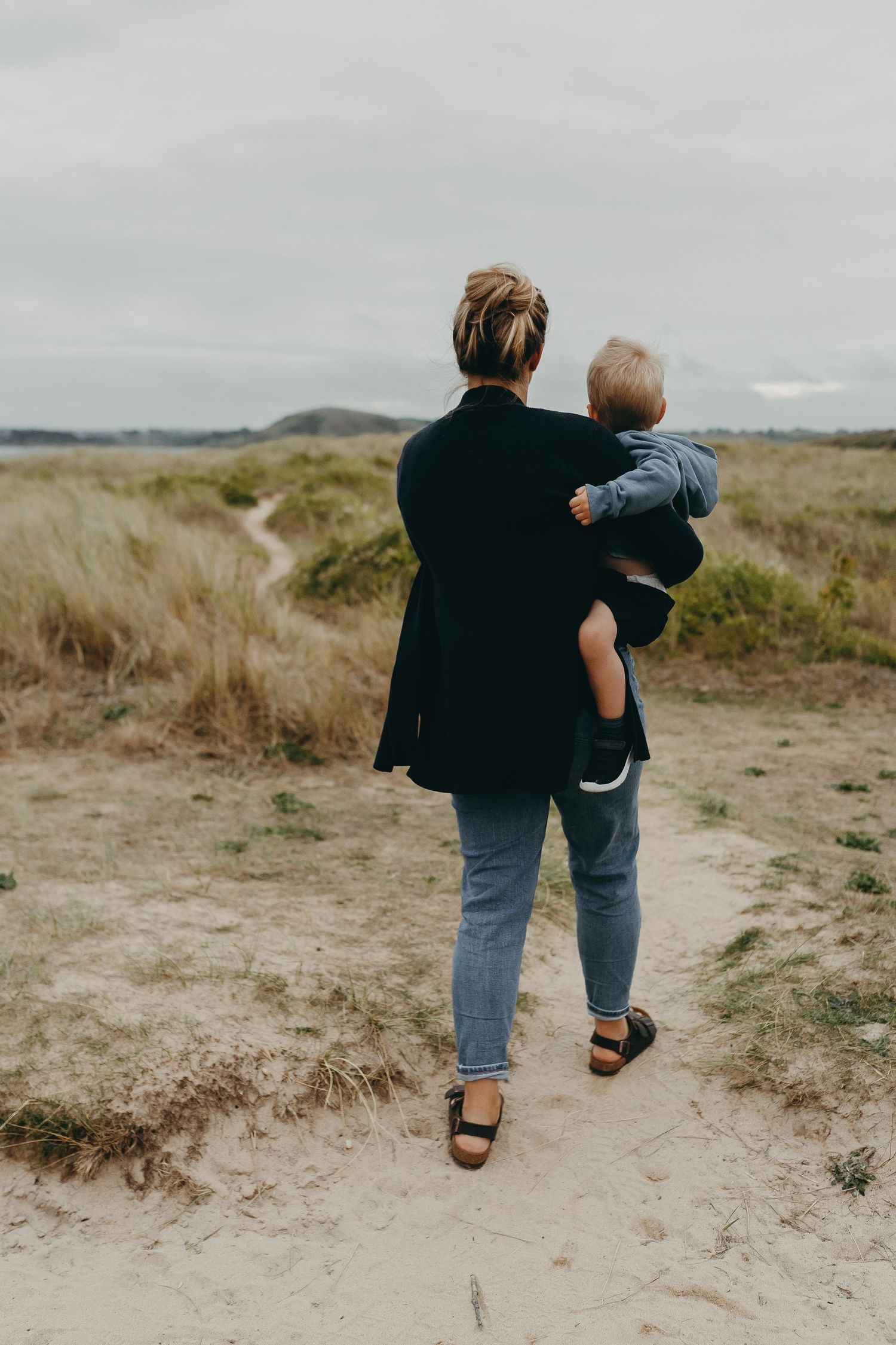 A person carrying a small child while walking along a coastal path on a cloudy day.