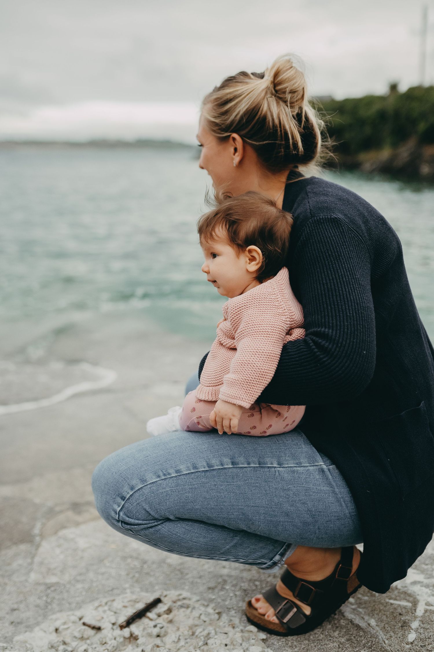 A tender seaside moment with parent and baby sitting on coastal rocks overlooking blue waters.