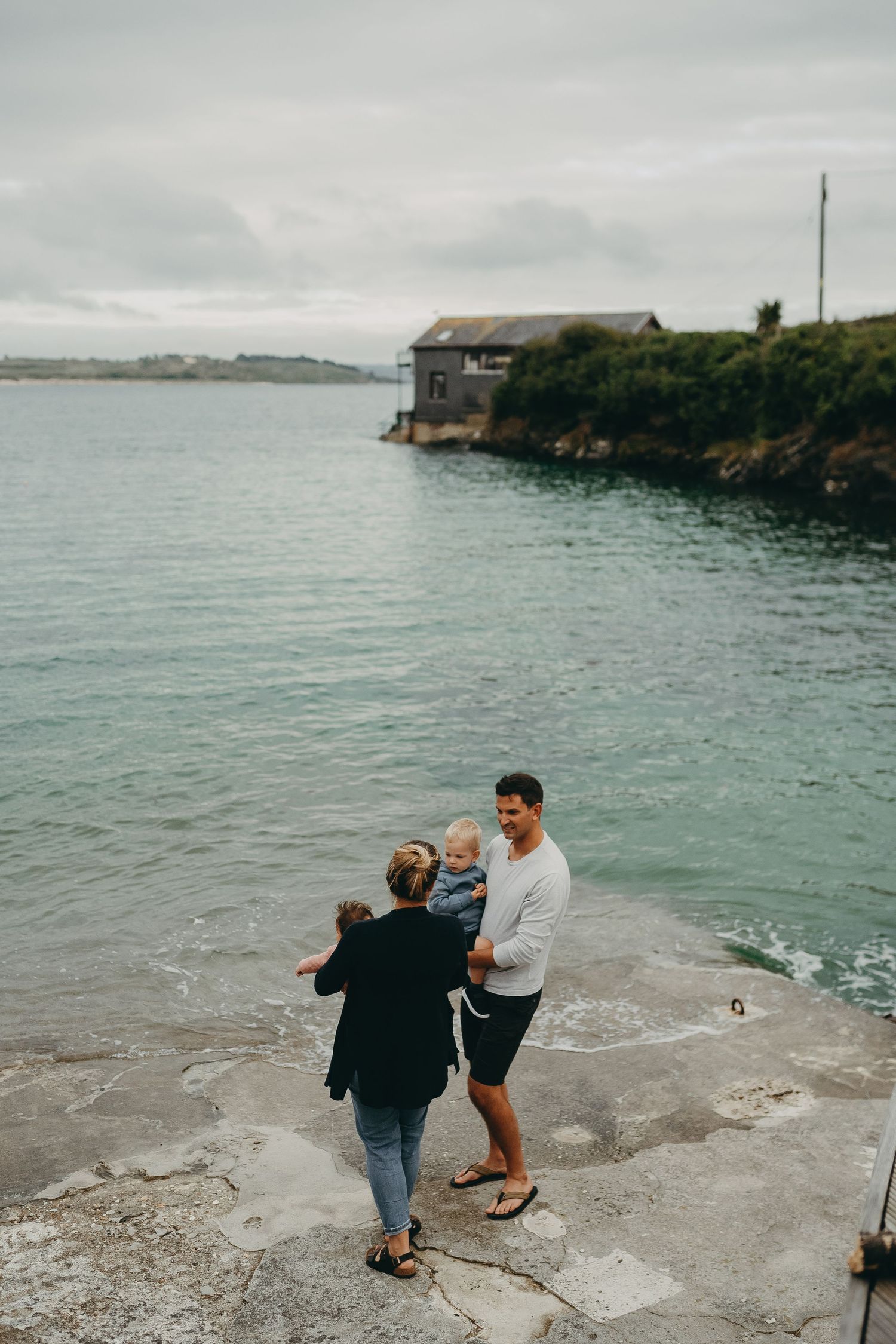 Three people stand on a rocky outcrop near a waterfront house on an overcast day.