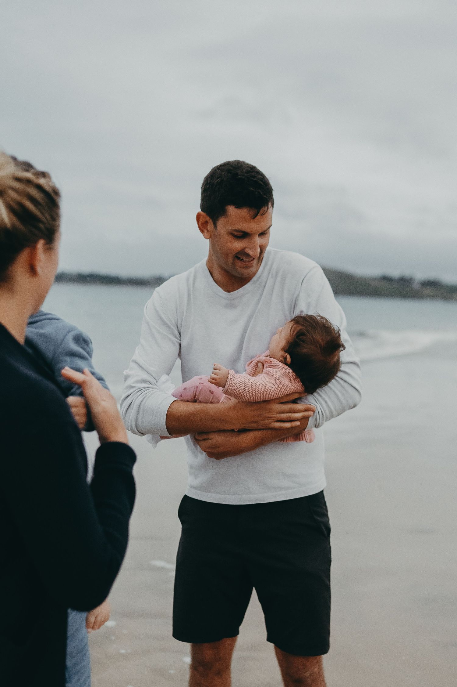 Family shares a tender moment with a baby at the beach on an overcast day.