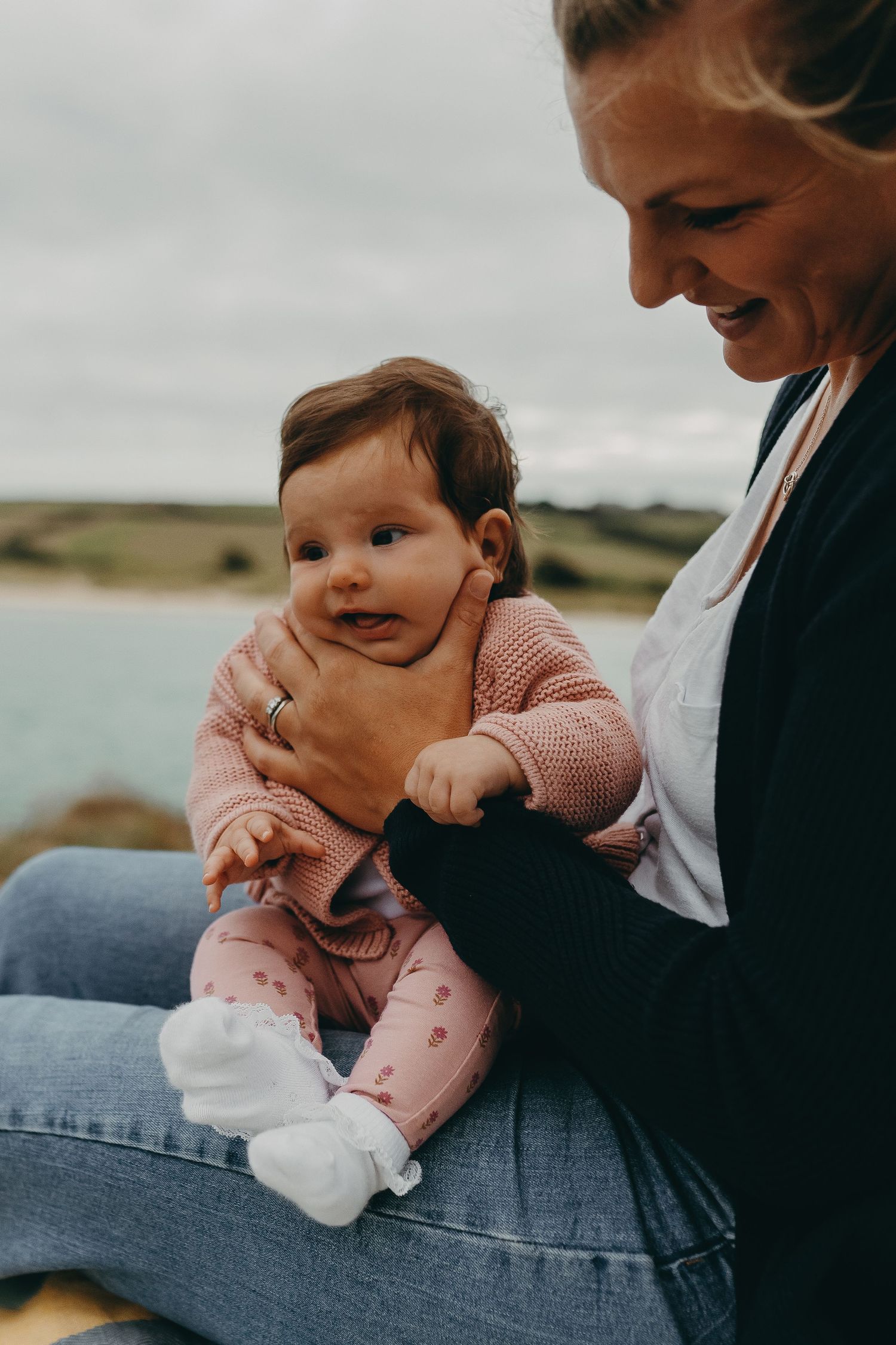 A sweet moment between parent and baby in pink sweater sitting by the ocean.