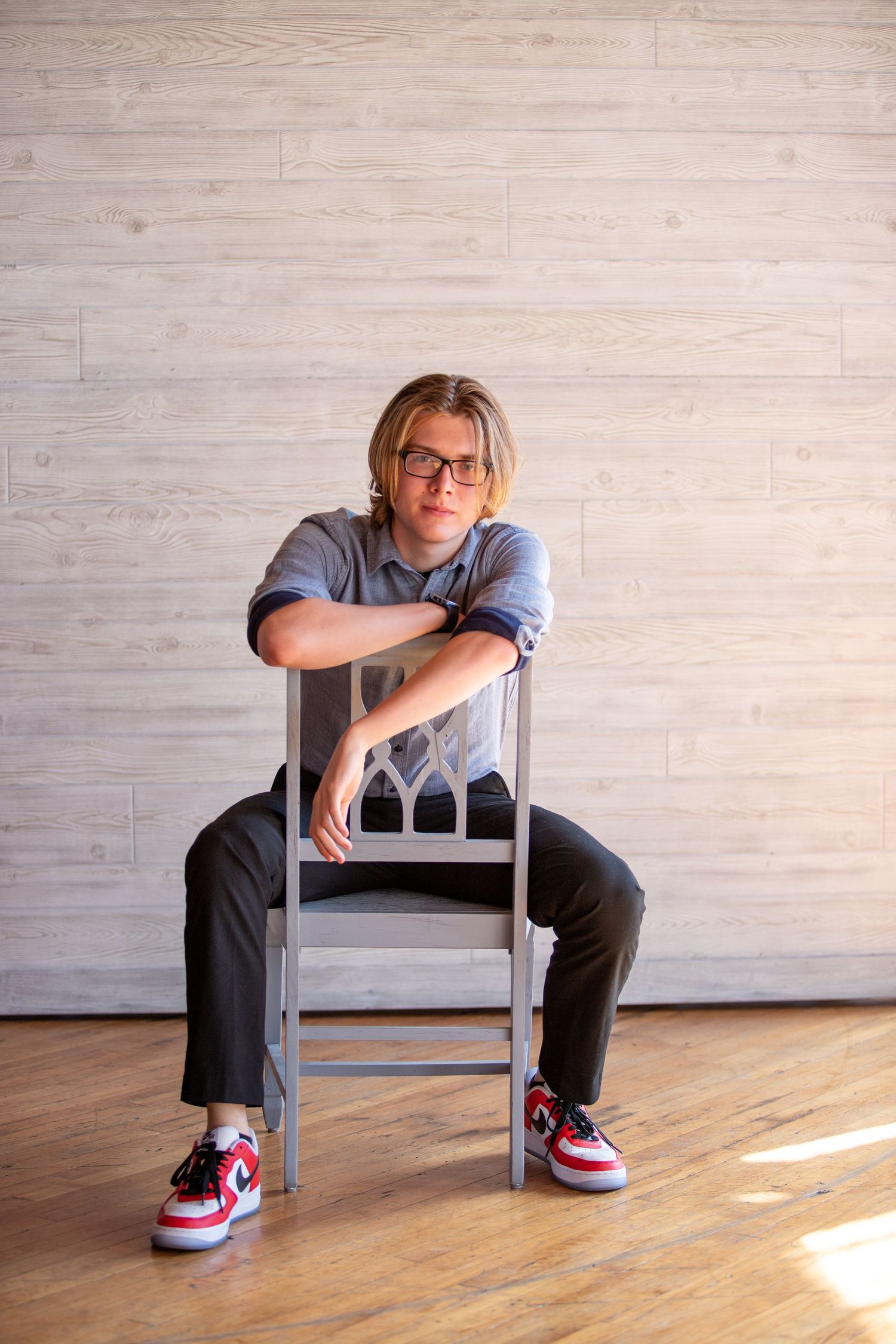 Teenage boy with glasses sitting backwards on a chair wearing Nike sneakers in a studio.