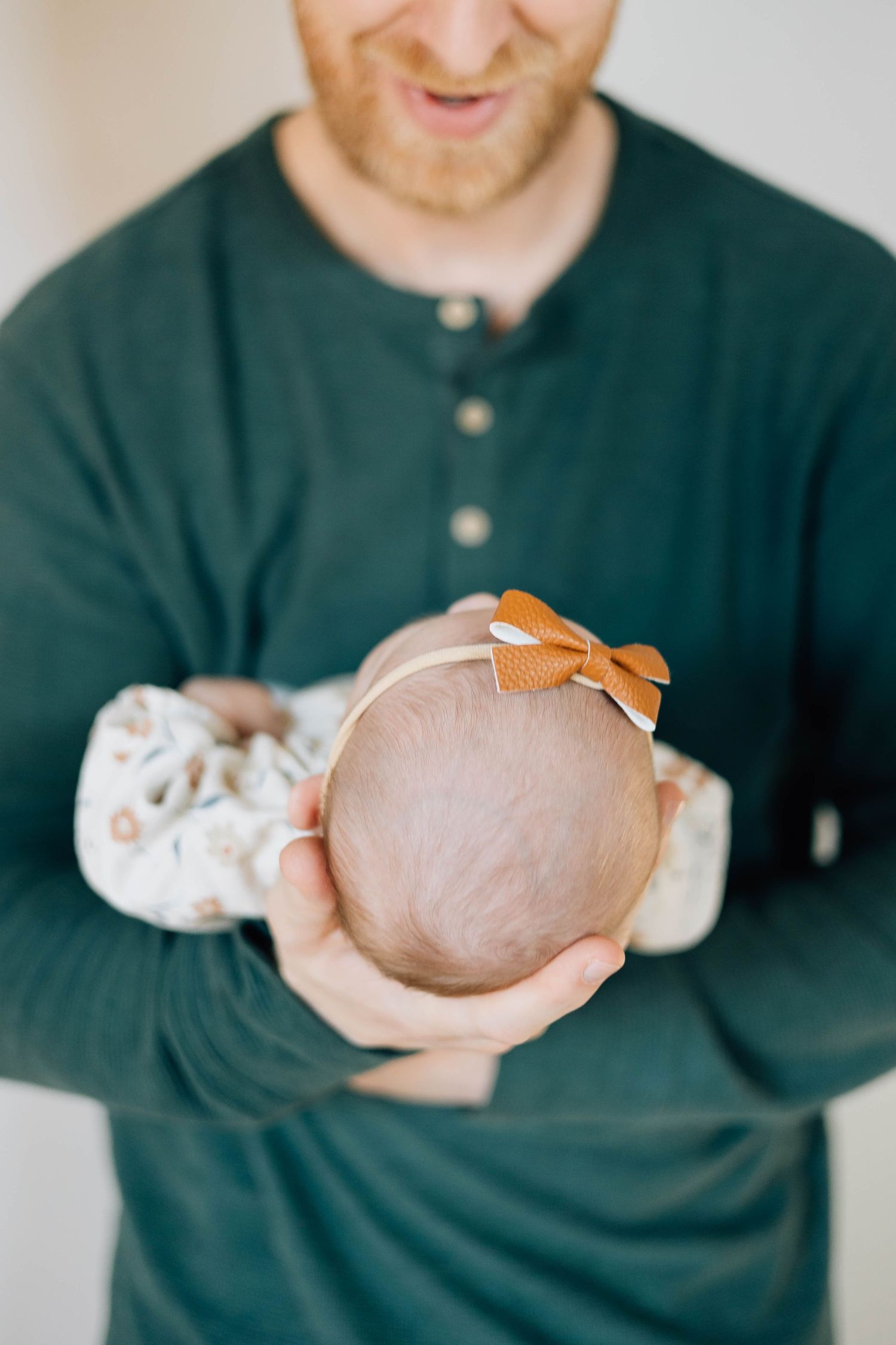 Father in a green henley shirt gently cradling his newborn baby girl wearing a brown bow headband.
