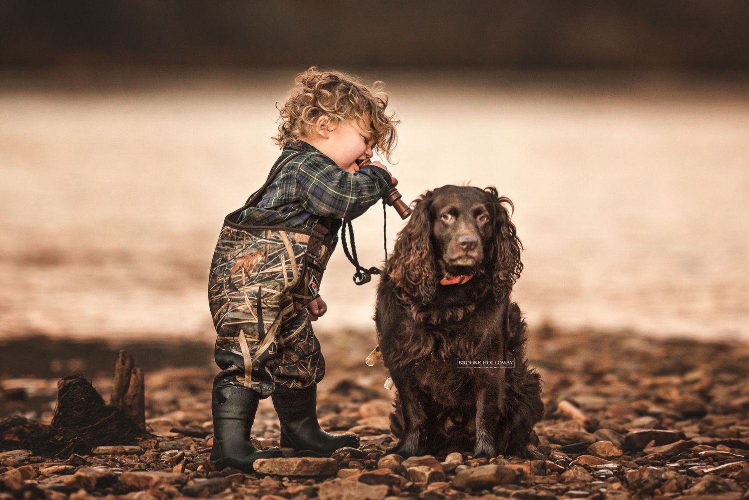 Small child in camo clothing walking with black dog through autumn leaves.