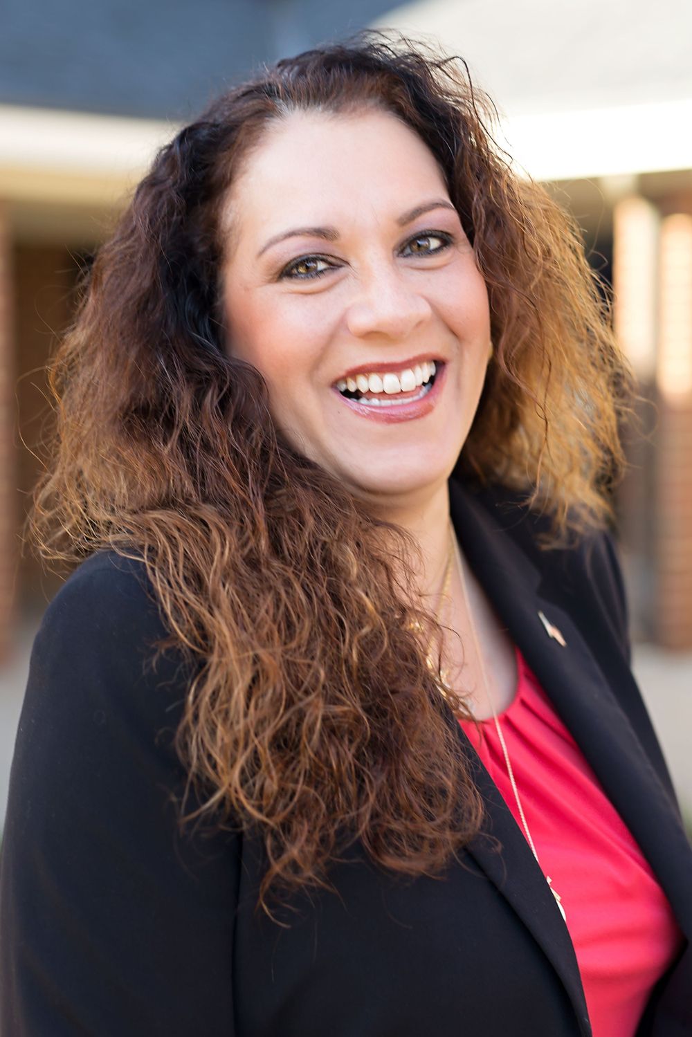 Professional headshot of person in black blazer and red top with curly hair.