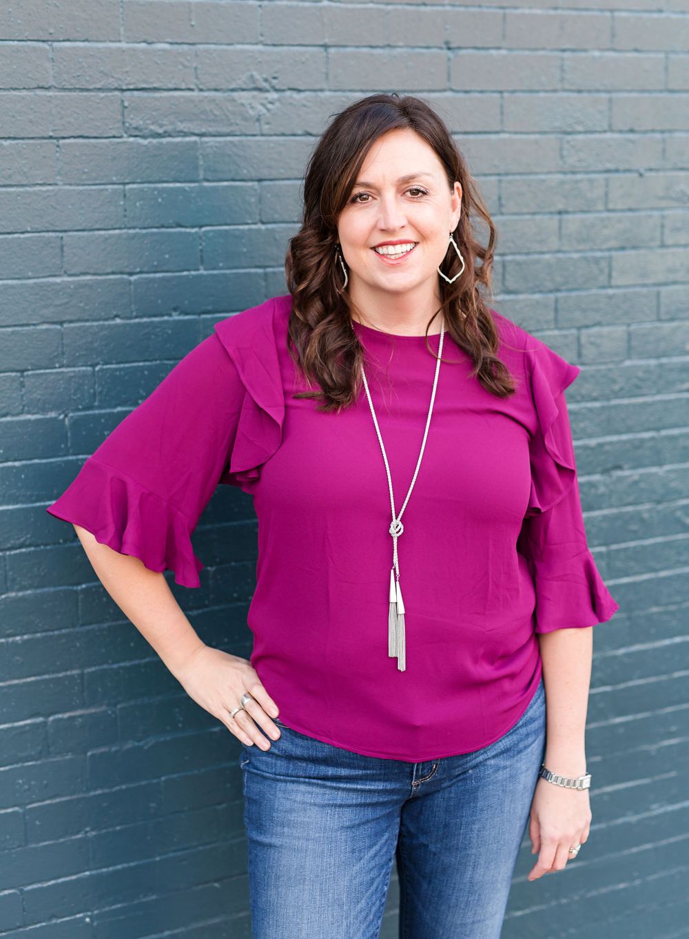 Woman in magenta ruffled sleeve top and jeans poses against blue brick wall.