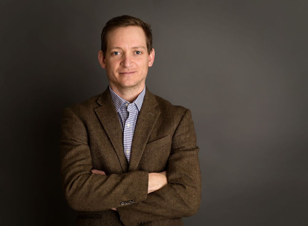 Professional headshot against dark background wearing brown blazer and checkered shirt.
