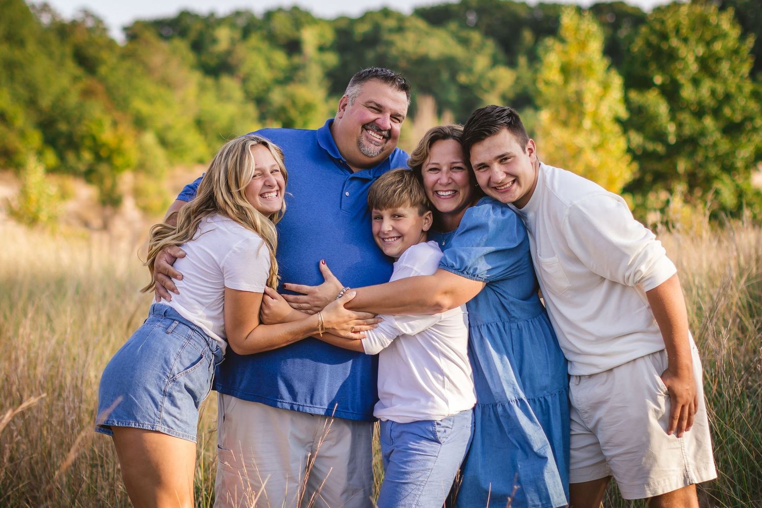 Happy family of five hugging and laughing together in a sunlit summer meadow.