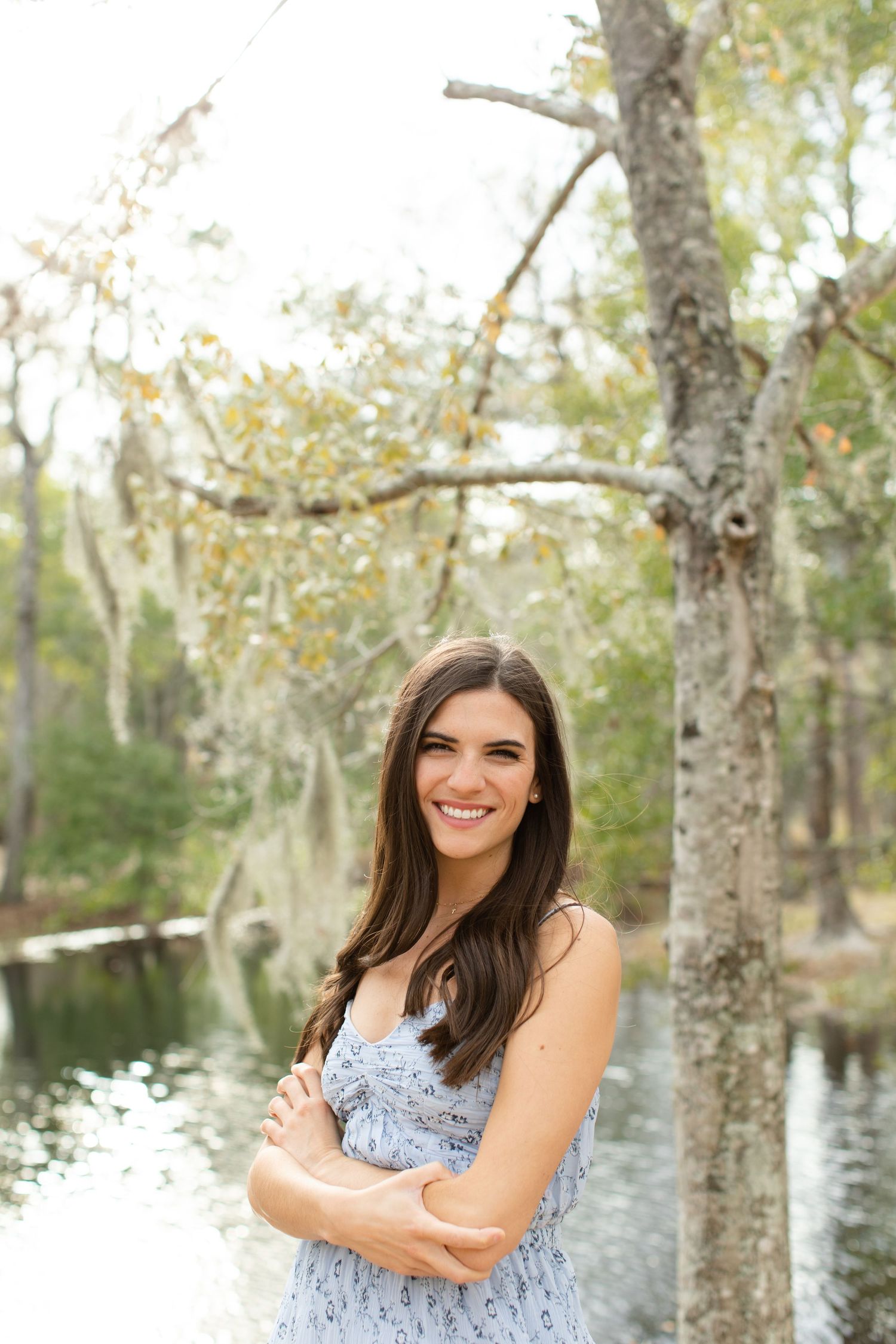Woman in blue dress poses confidently by water with autumn trees in background.