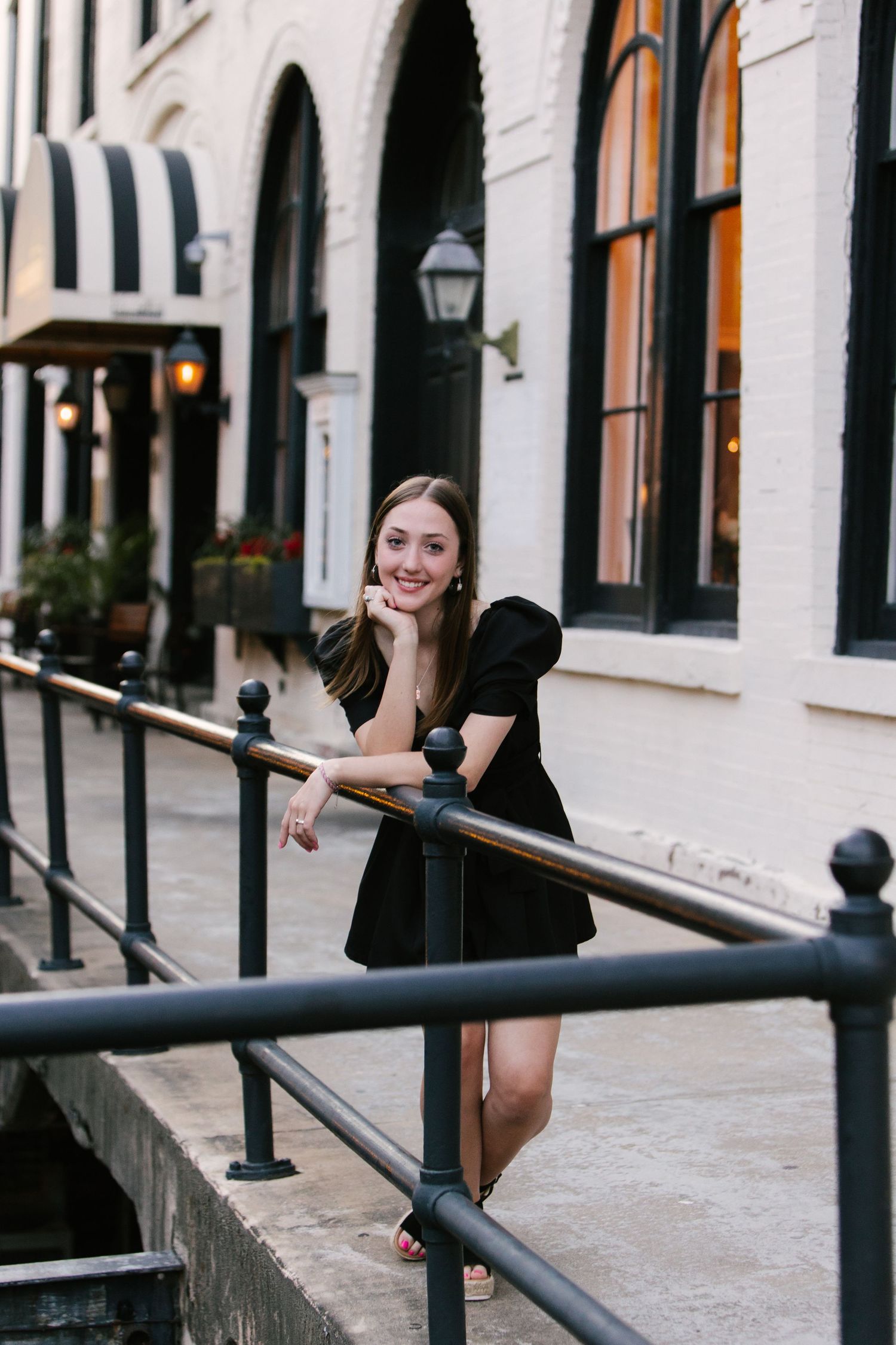 Woman in a black dress leaning on railing outside historic building with arched windows.